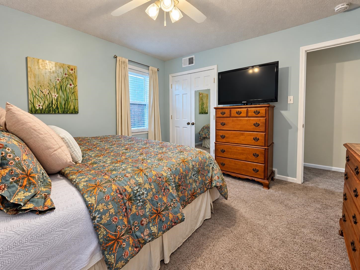 Bedroom with a floral bedspread, wooden dresser and television, and a window with cream curtains.