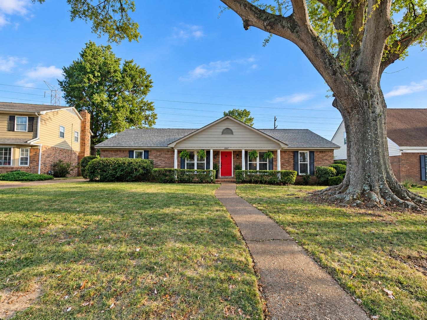 Single-story brick house with a red front door, front lawn, and large shade tree.