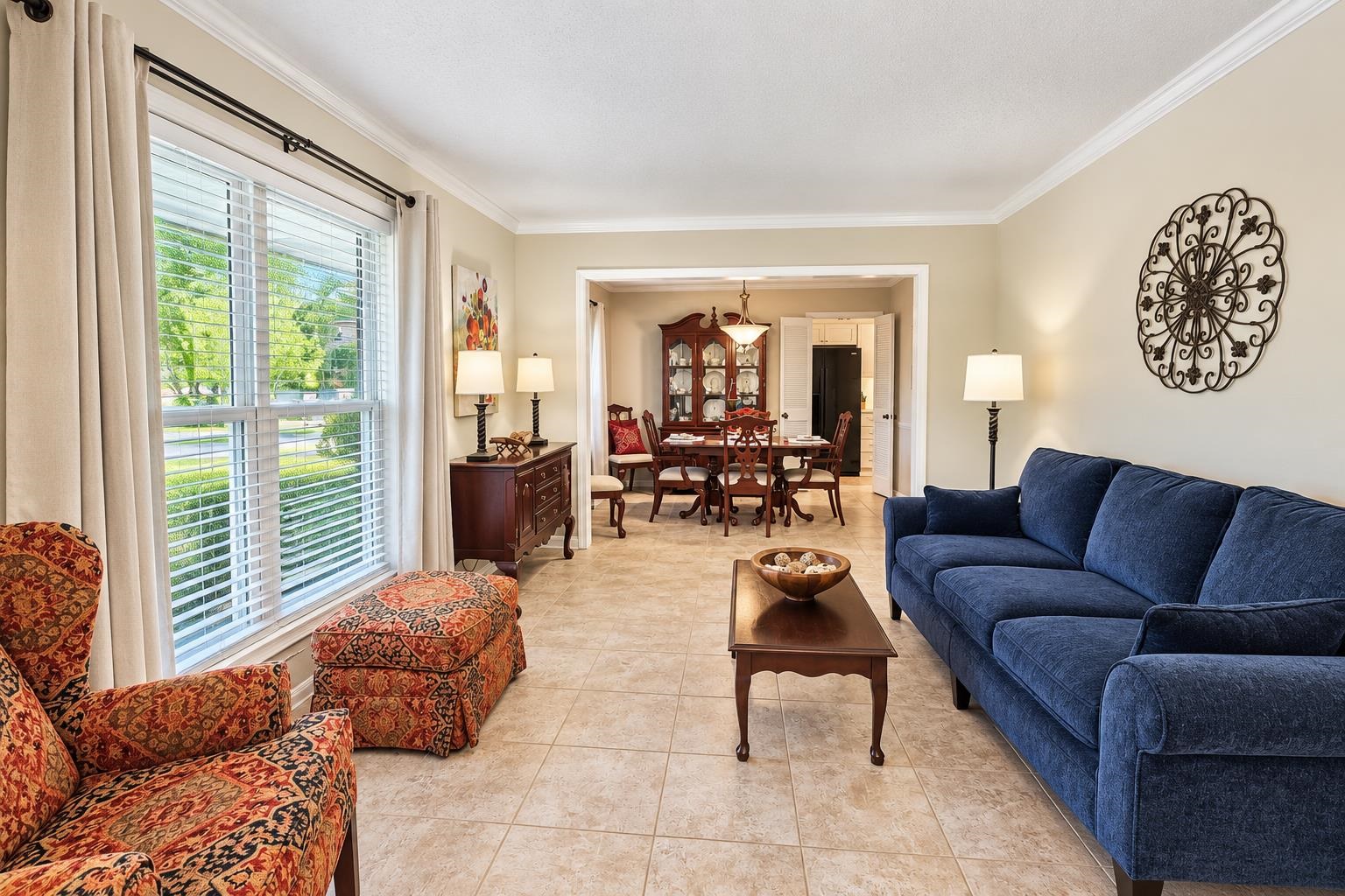 Living room with blue sofa and patterned chairs, opening to a dining room with a wooden table.