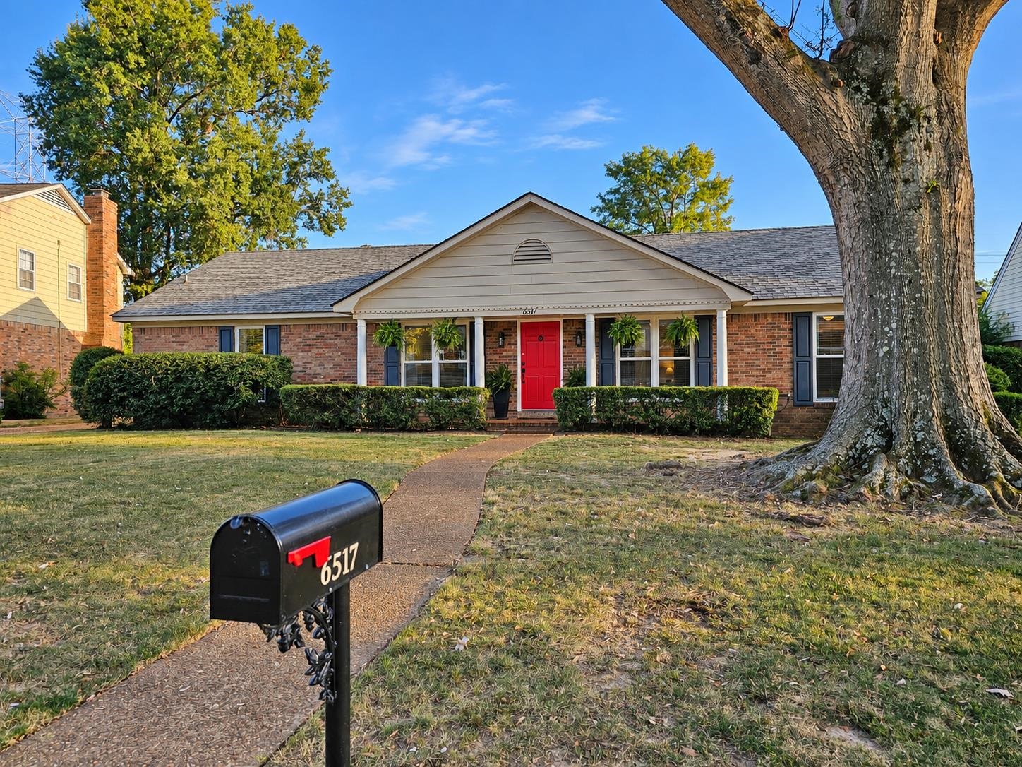 Brick ranch house with a bright red front door, curved walkway, and large shade tree.