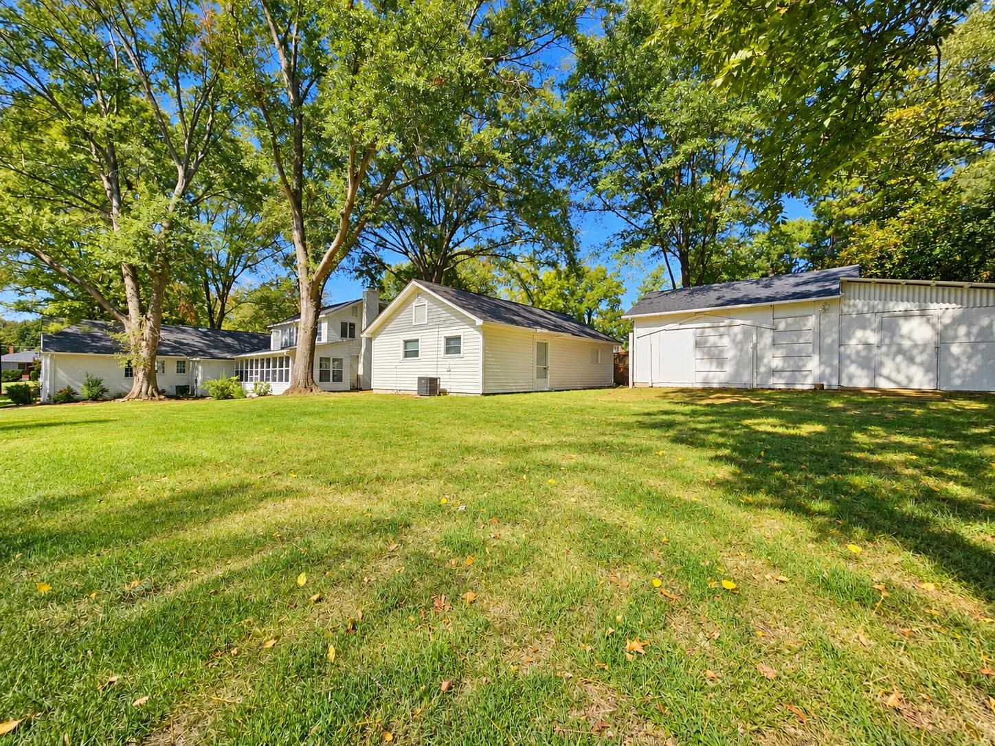 Light-colored home and detached garage beyond a broad lawn shaded by mature trees.