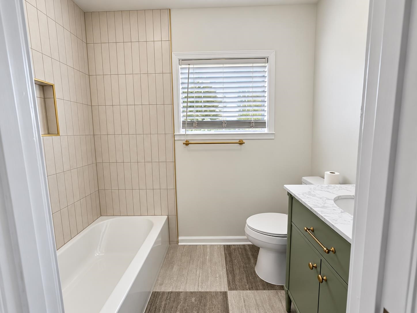 Bathroom with beige tiled tub surround, green vanity, brass hardware, and a window with blinds.