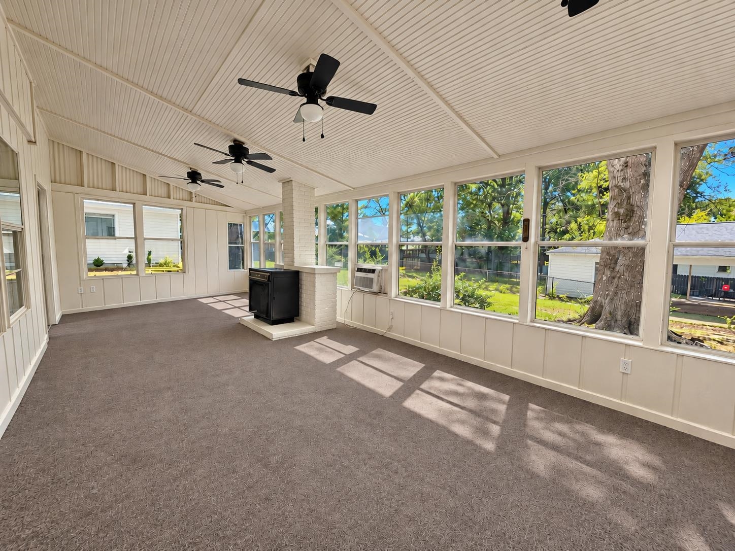 Enclosed sunroom with broad backyard-facing windows, three ceiling fans, carpet, and a black stove on a brick hearth.