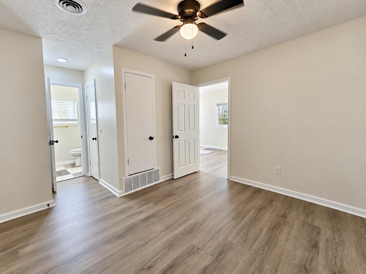 Empty room with wood-look flooring and a ceiling fan, opening to a bathroom and adjoining room.