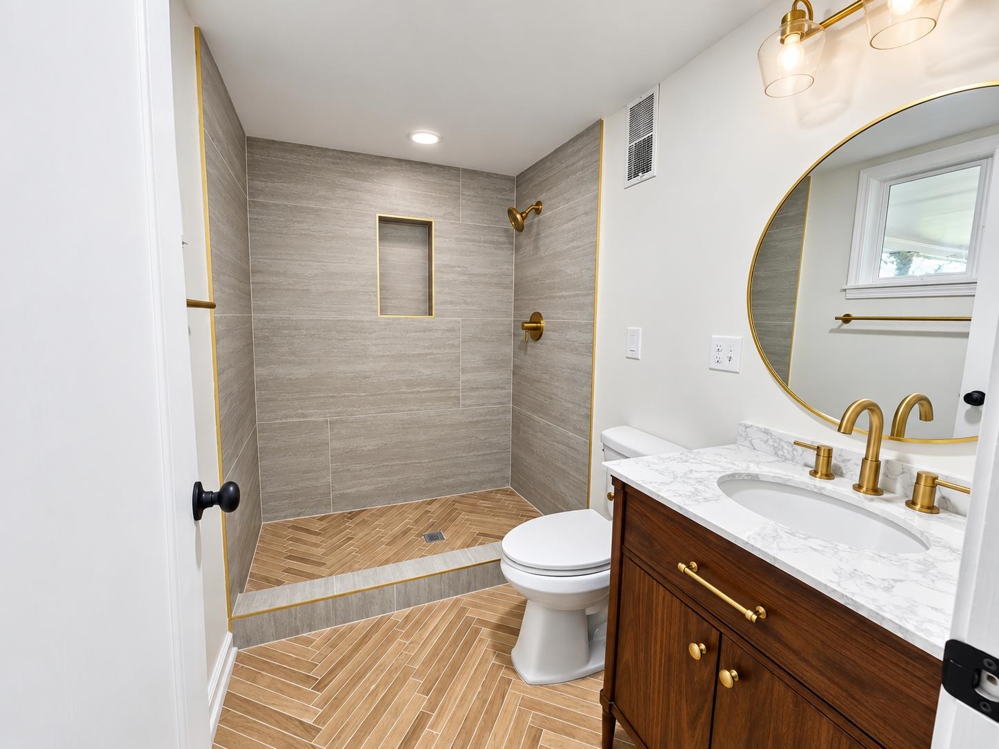 Renovated bathroom with a tiled walk-in shower, herringbone floor, dark wood vanity, and round mirror.