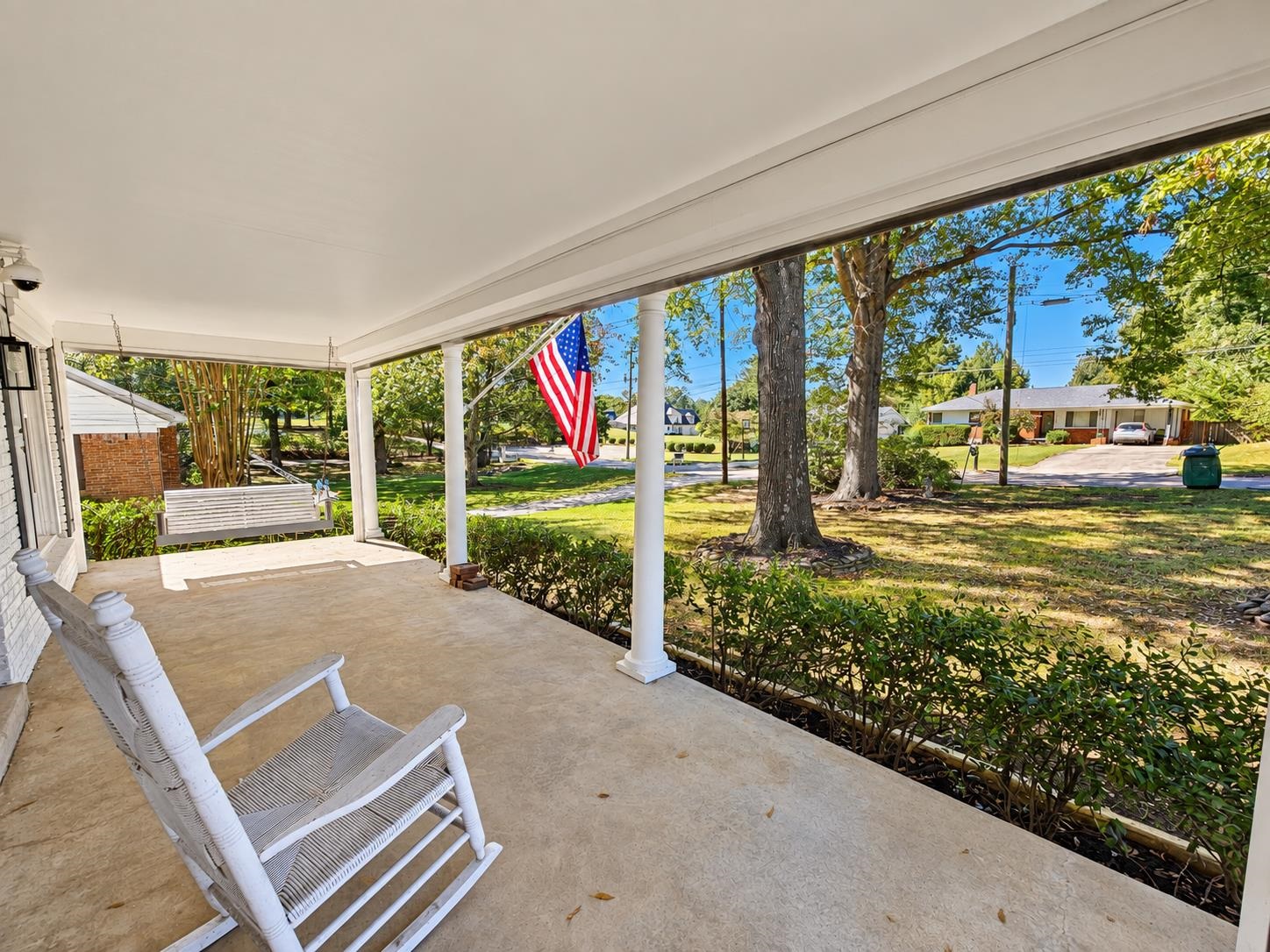 Covered front porch with white rocking chairs and an American flag overlooking a leafy residential street.