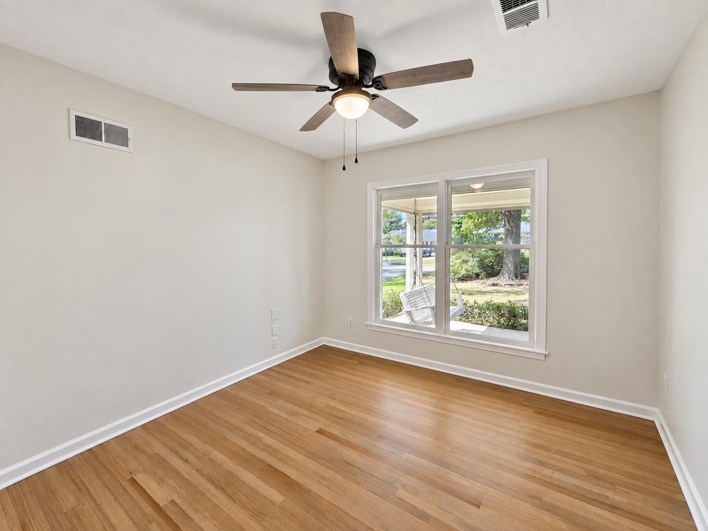 Empty room with hardwood floors, a ceiling fan, and a large window overlooking a leafy yard.