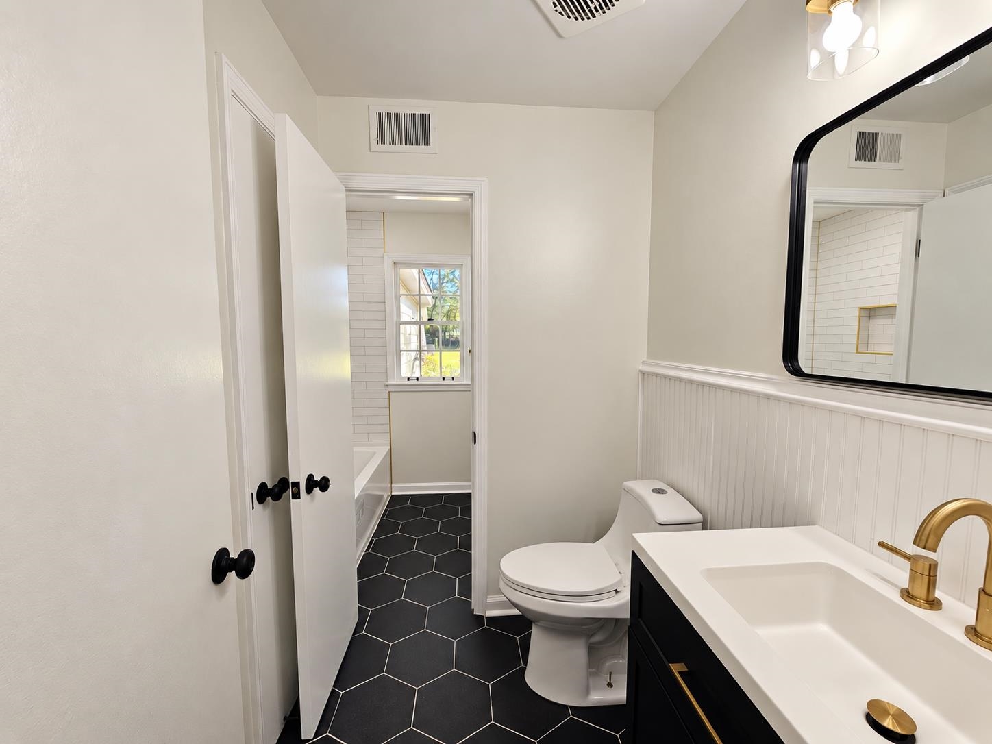 Bathroom with black hexagonal floor tile, a black vanity, brass faucet, and tiled bathing area beyond.