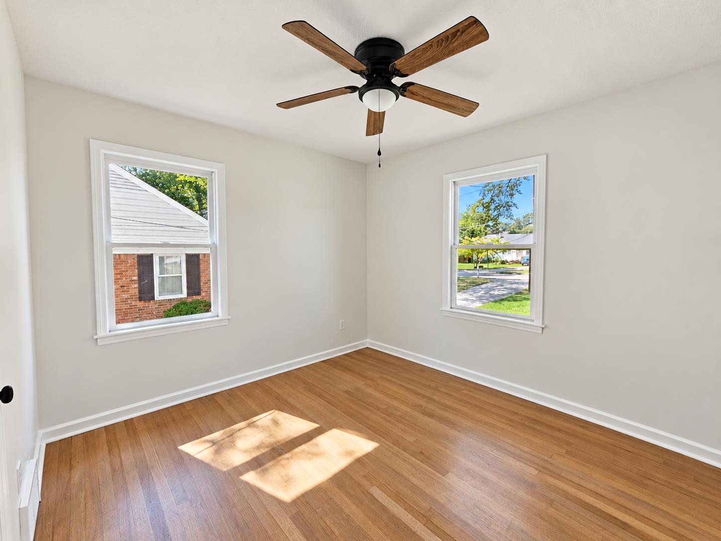 Empty room with hardwood floors, two windows, and a wood-blade ceiling fan.