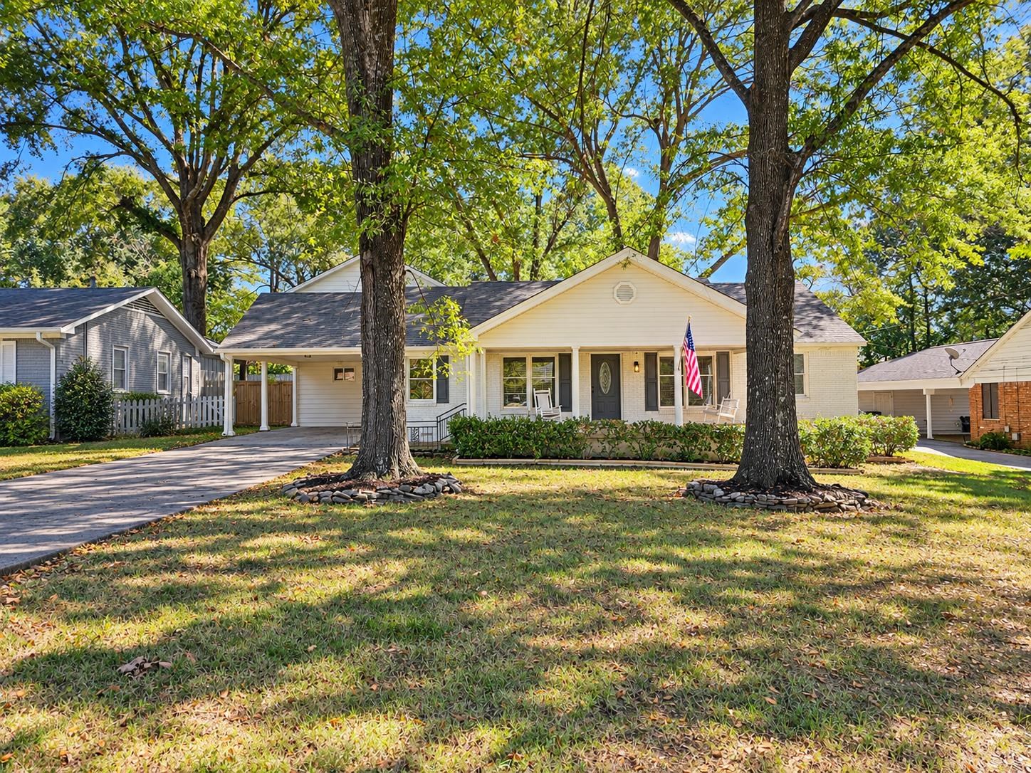 Single-story home with a covered porch, American flag, and mature trees shading the front lawn.