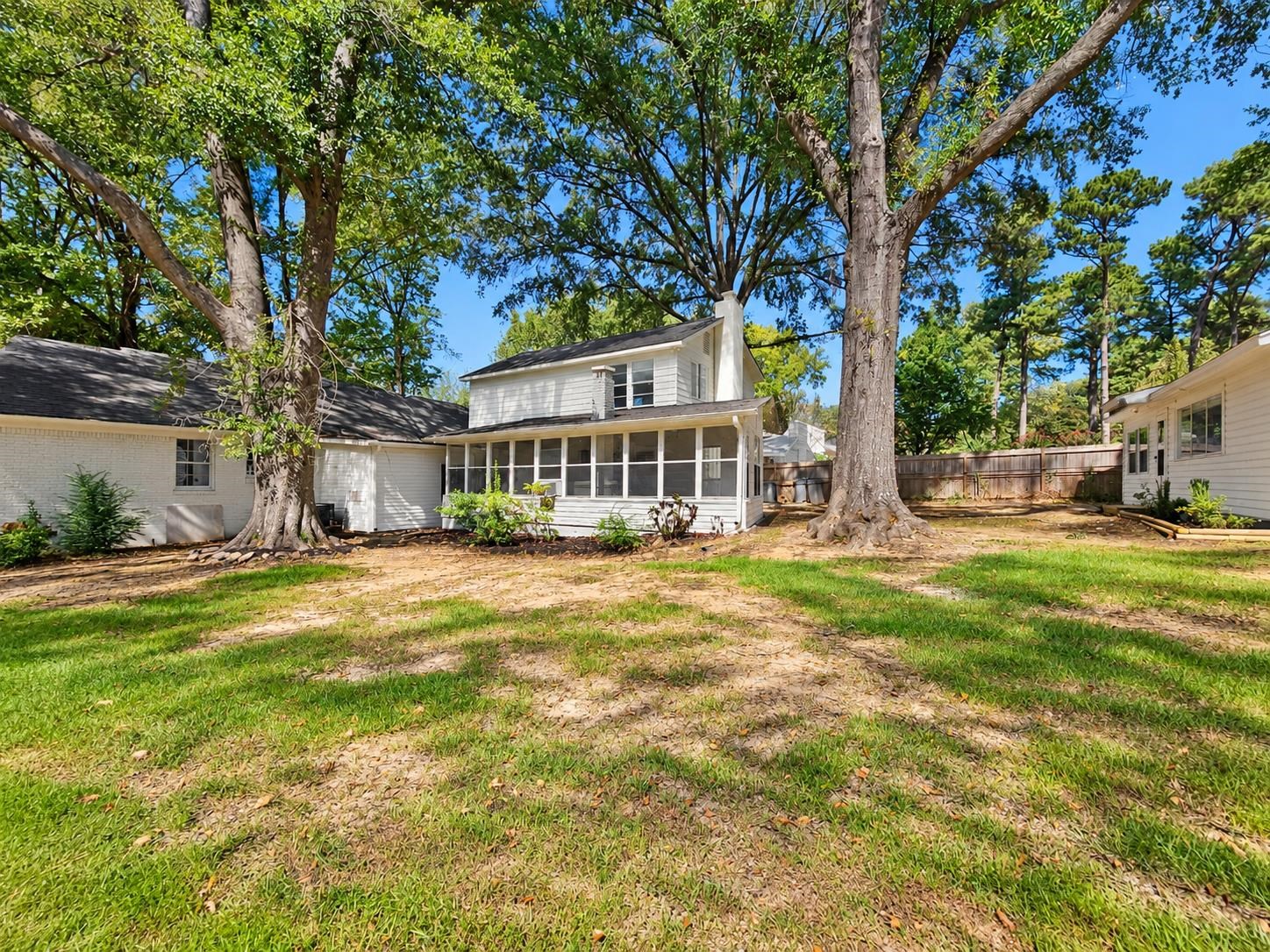 White house with a screened rear porch beneath mature trees, opening onto a fenced lawn.
