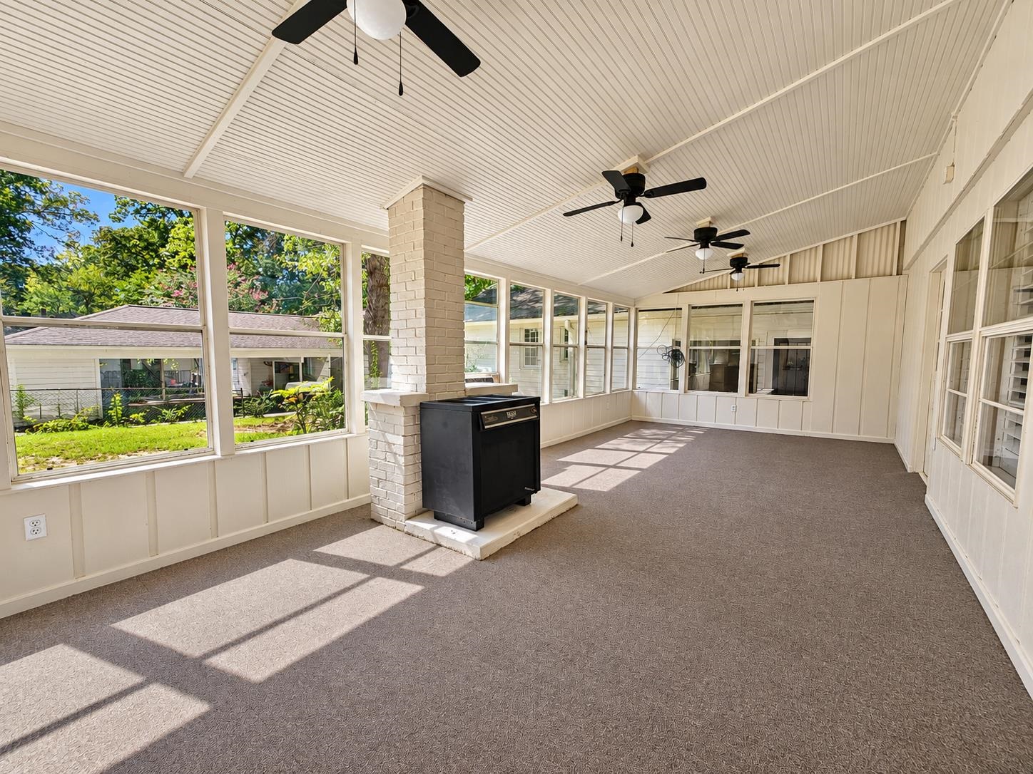 Enclosed sunroom with large yard-facing windows, carpet, three ceiling fans, and a black stove.