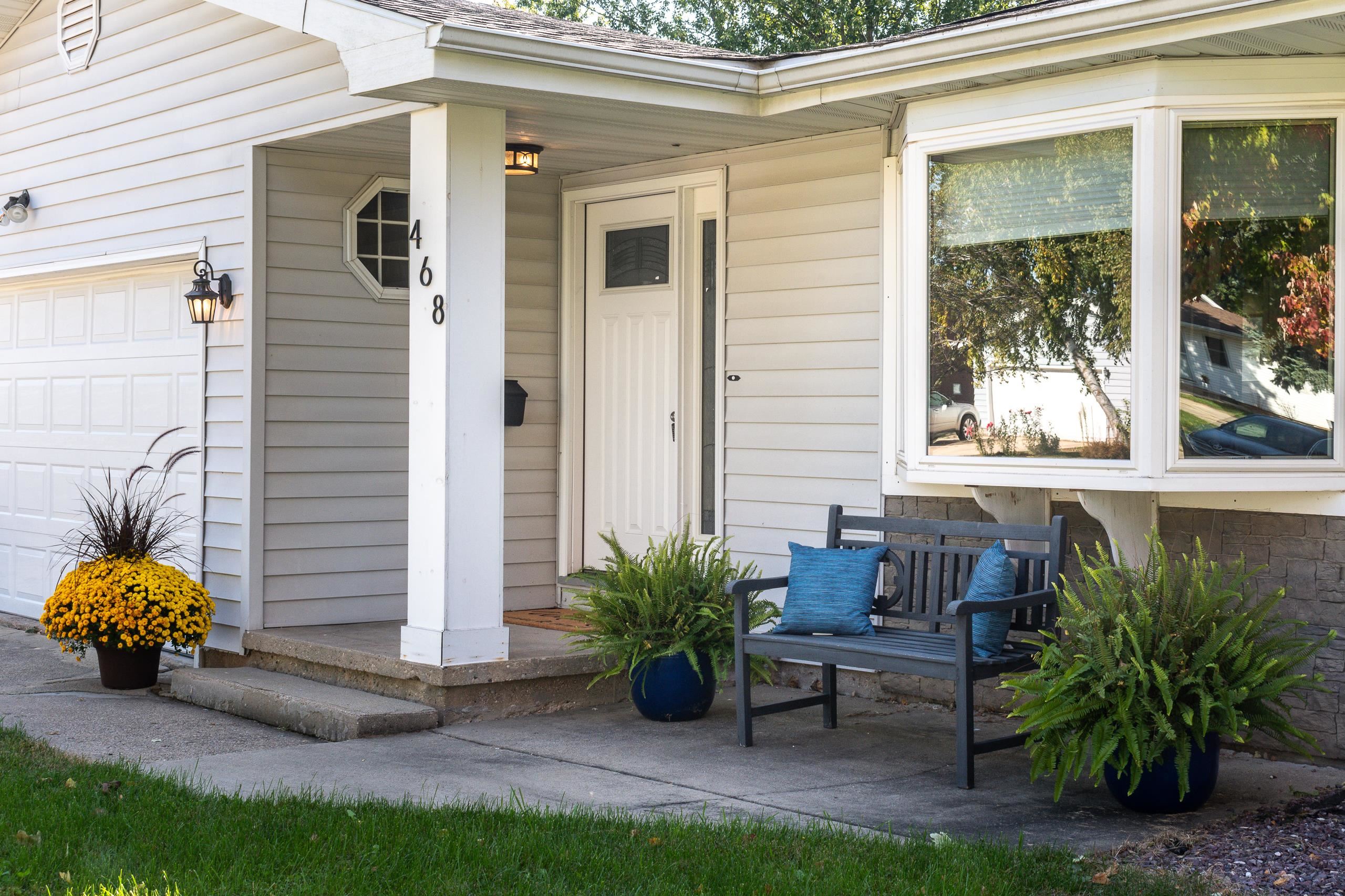 Home entrance with a covered porch, white door, and bench surrounded by potted plants.