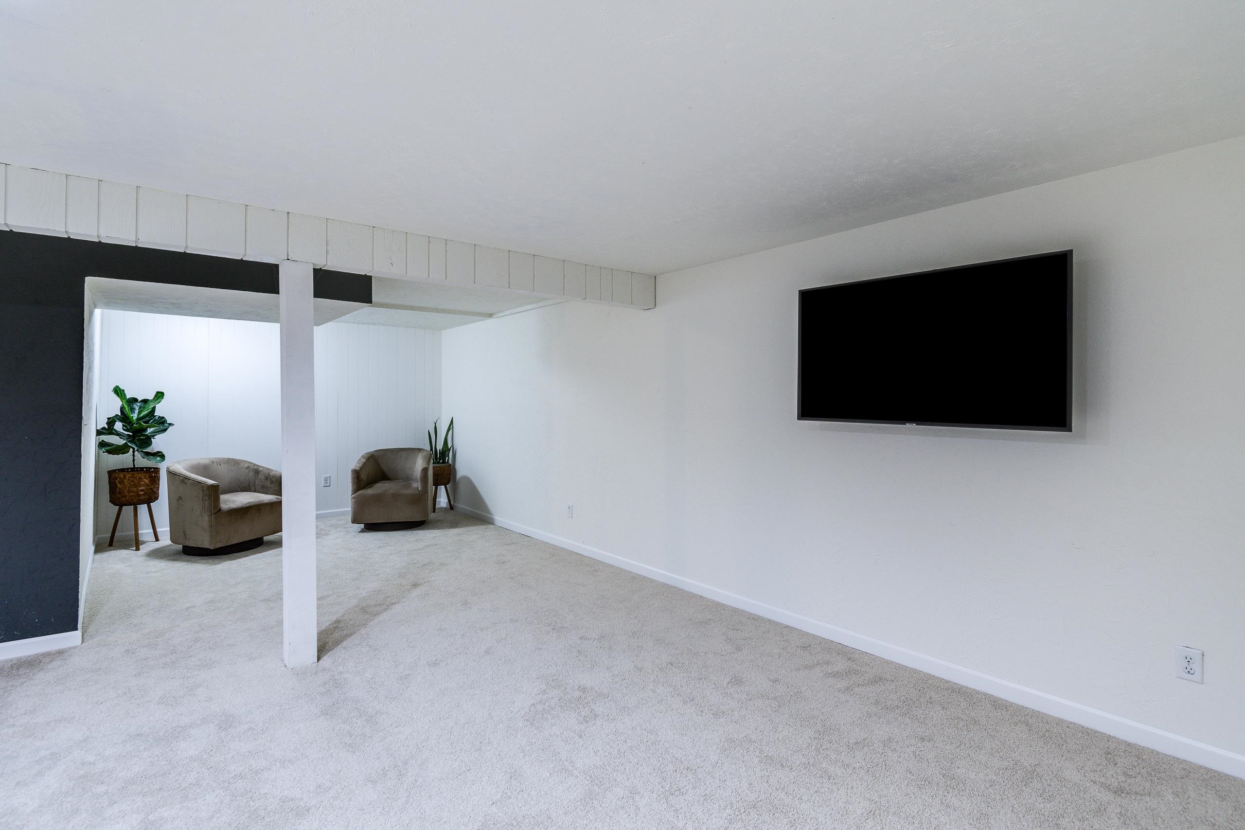 Carpeted living area with a wall-mounted television and a seating nook with chairs and plants.