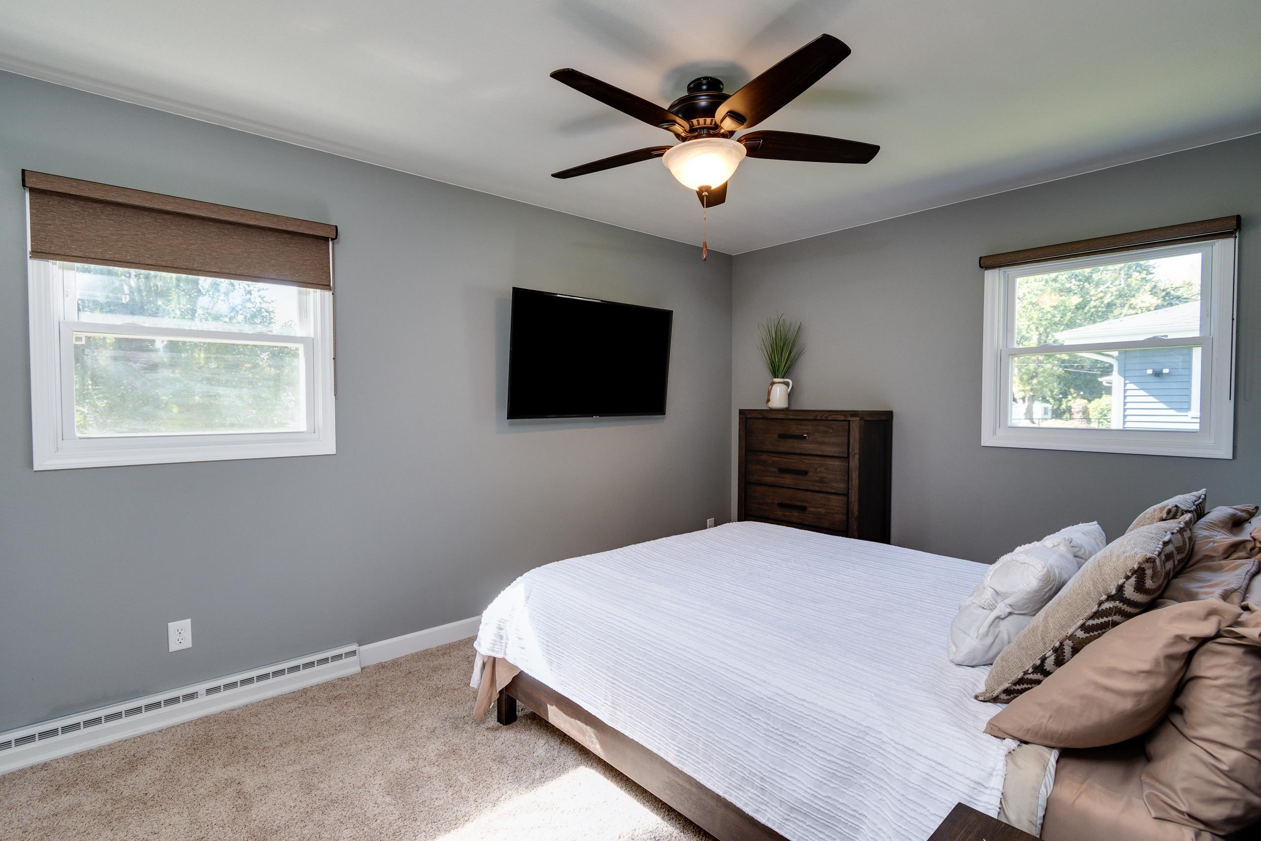 Bedroom with a white bed, gray walls, two windows, and a dark wood dresser beneath a ceiling fan.