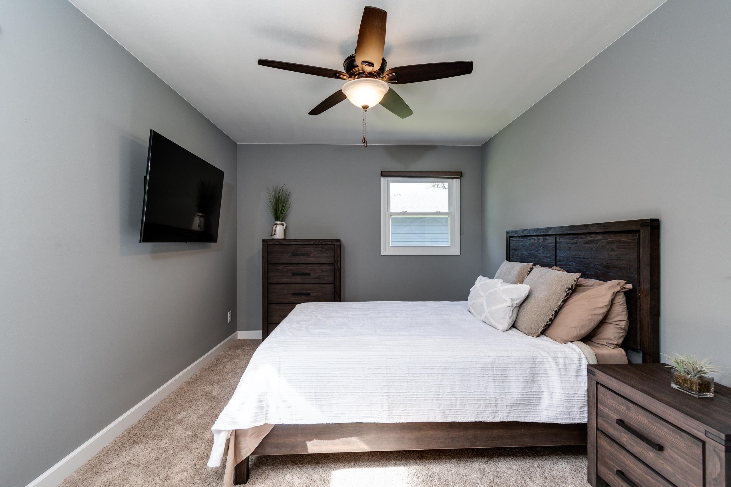 Furnished bedroom with a dark wood bed, white bedding, wall-mounted TV, and ceiling fan.