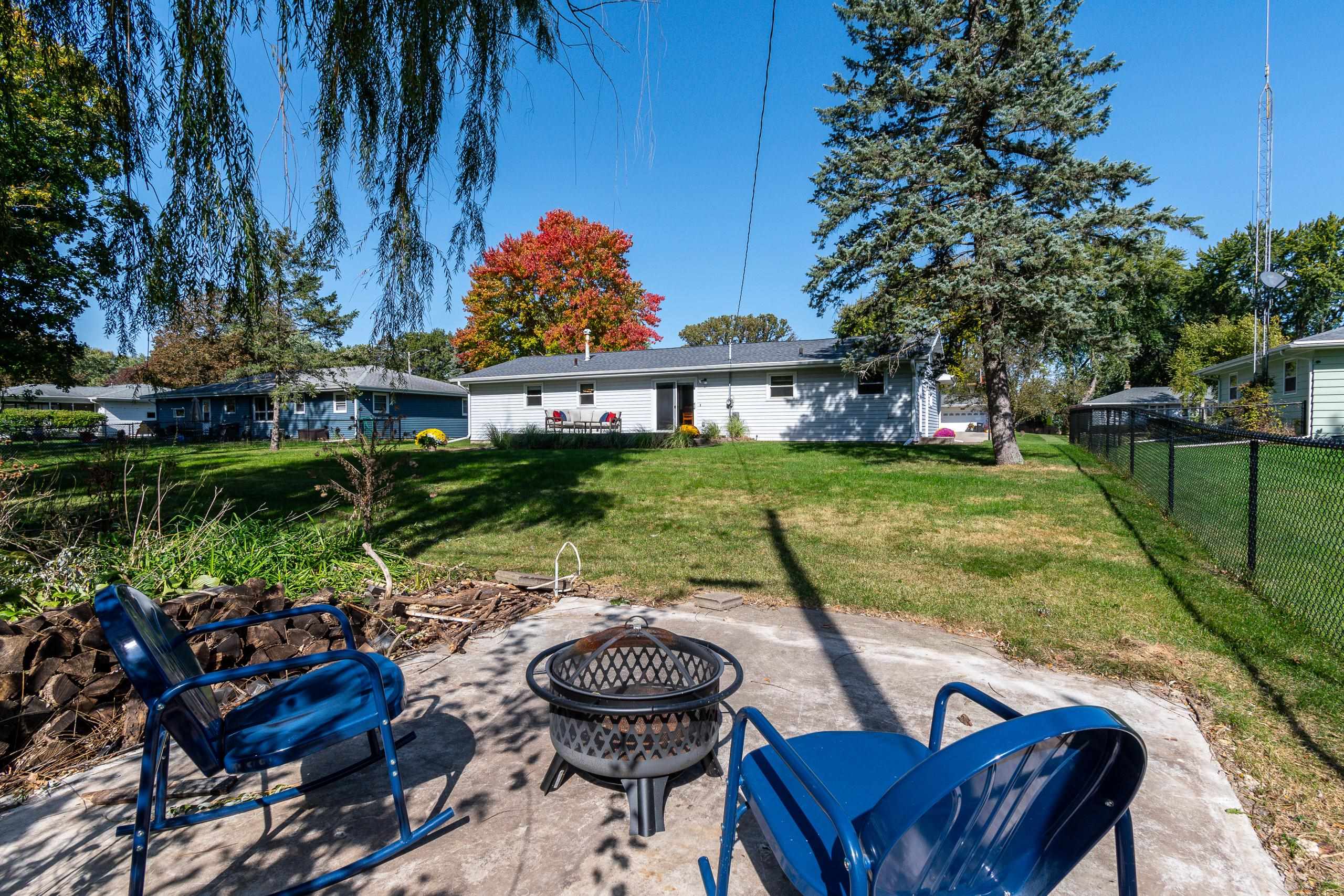 Backyard patio with blue chairs and fire pit facing a single-story home and fenced lawn.