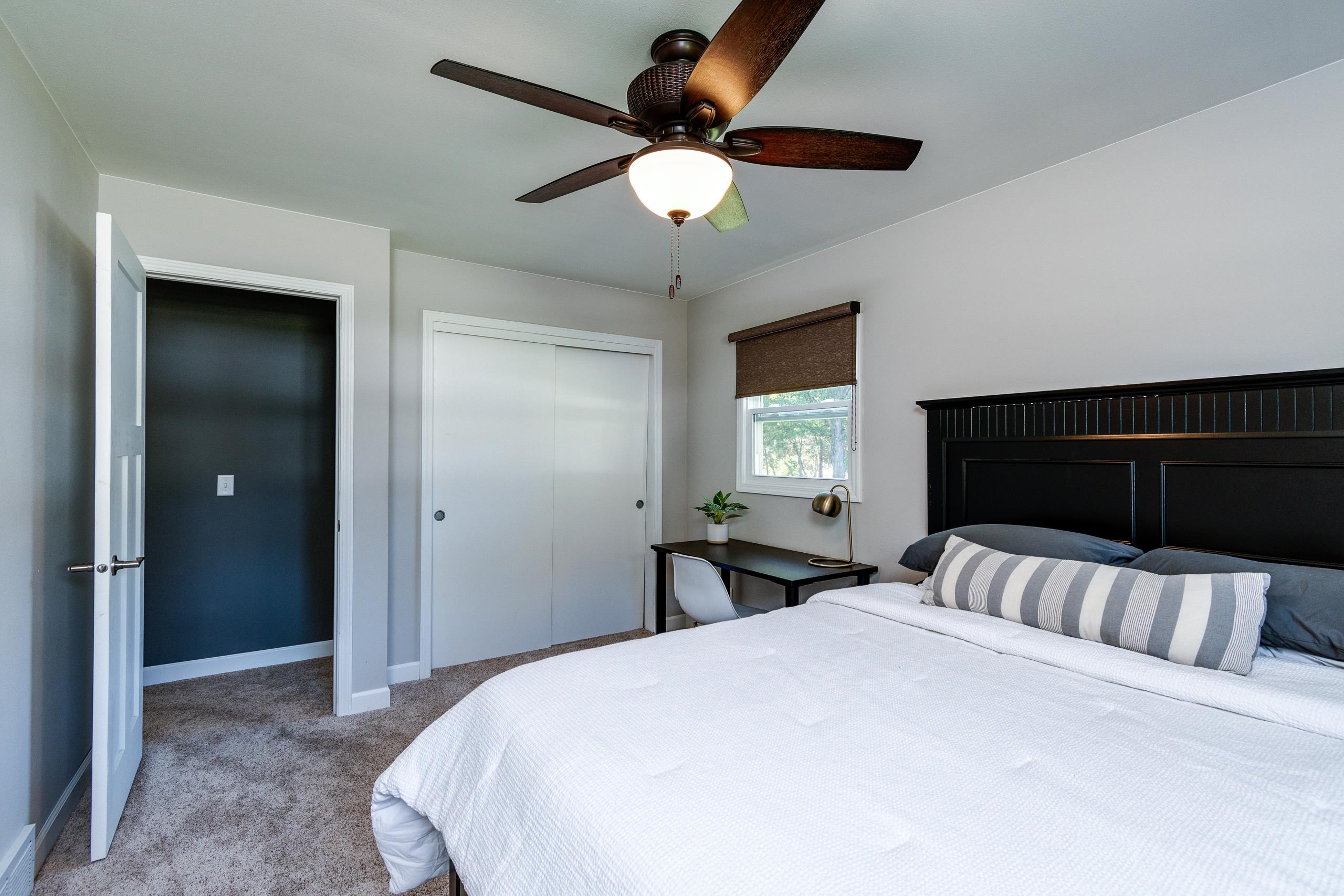 Bedroom with a dark paneled headboard, striped pillow, ceiling fan, and desk beside the window.