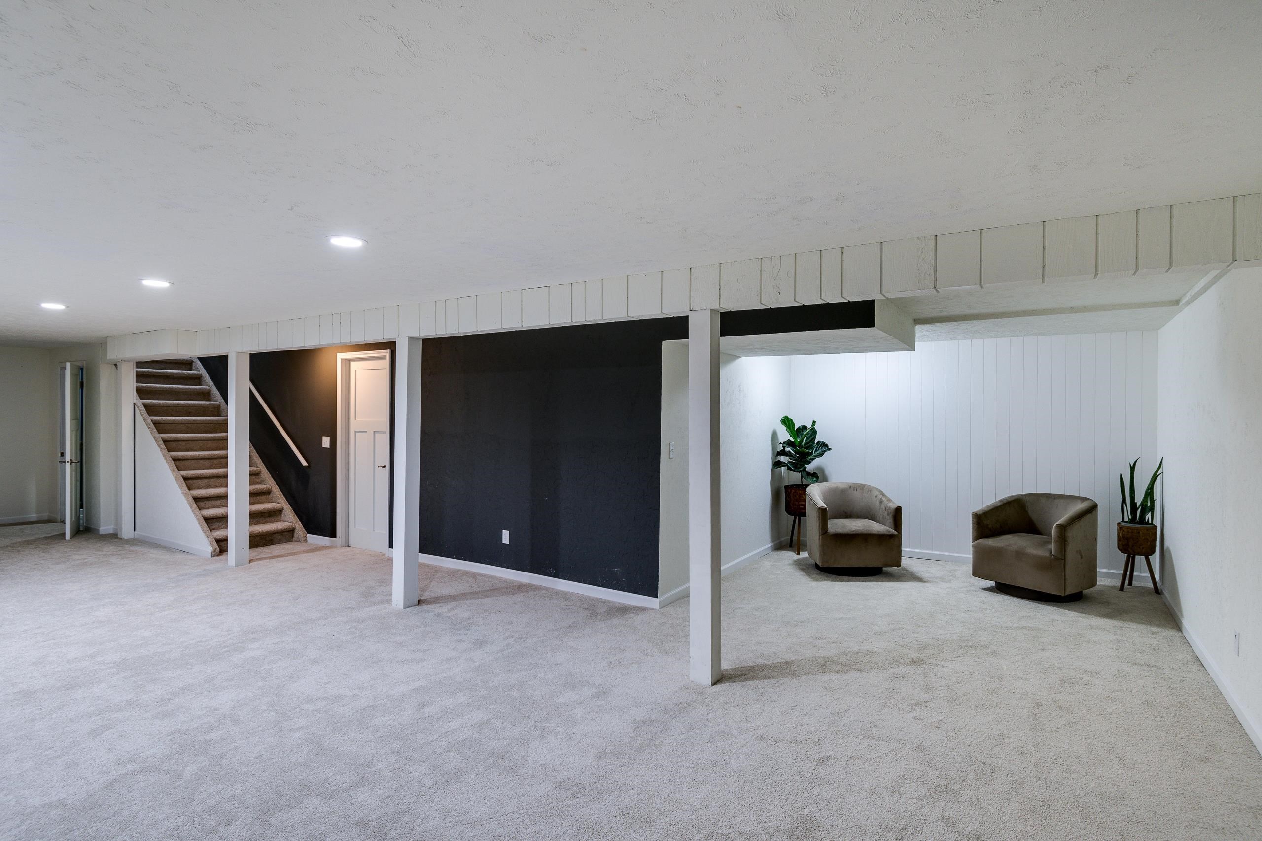 Finished basement with carpeted stairs, a dark accent wall, and two armchairs in a seating area.
