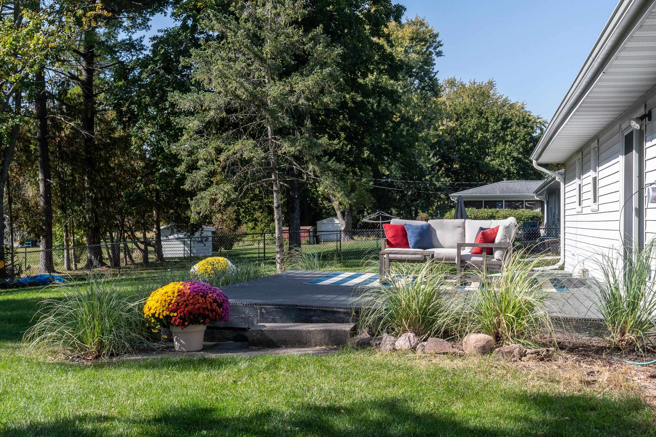 Backyard patio with a cushioned outdoor sofa, potted mums, ornamental grasses, and mature trees.