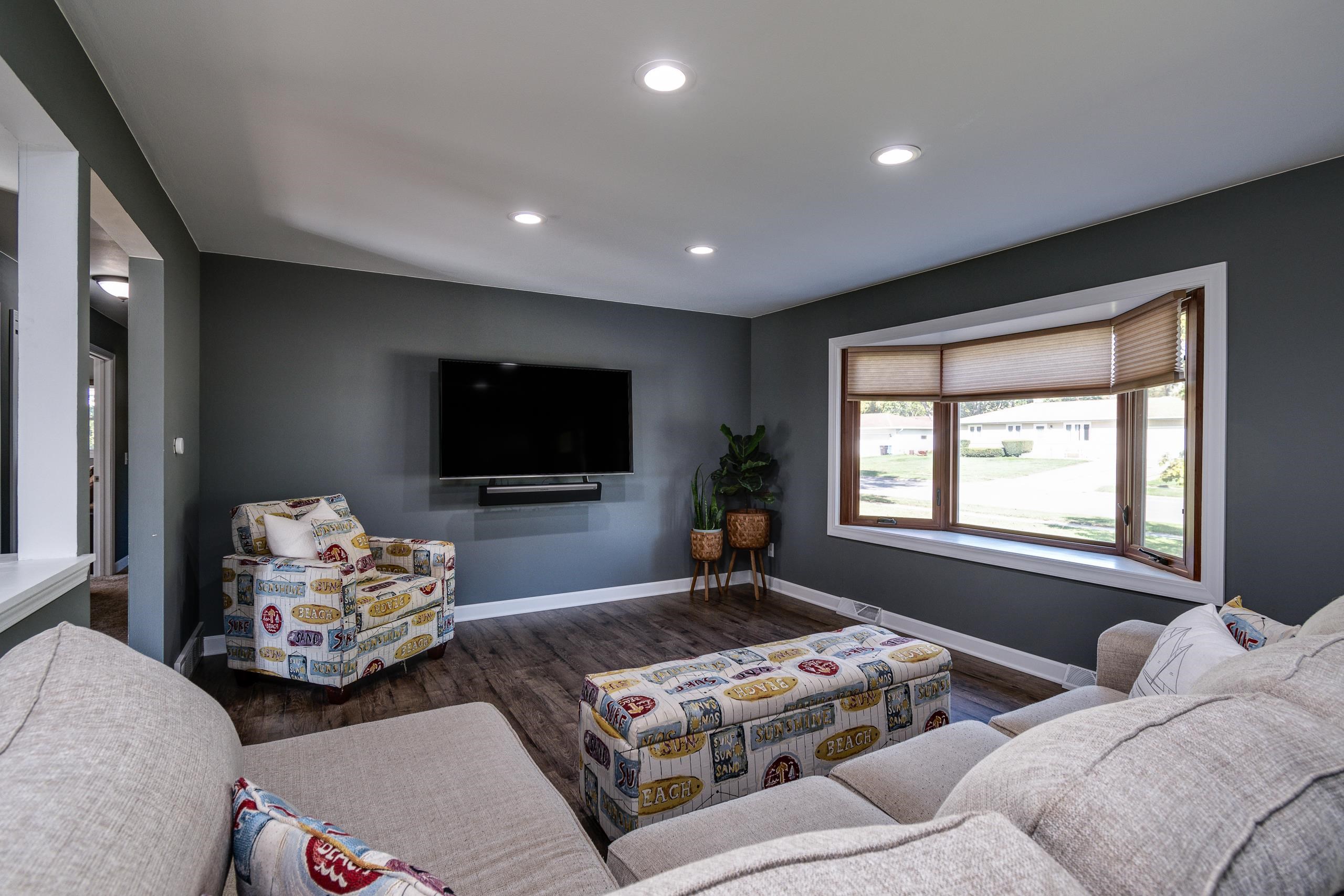 Living room with a wall-mounted TV, patterned chair and ottoman, and a large bay window.