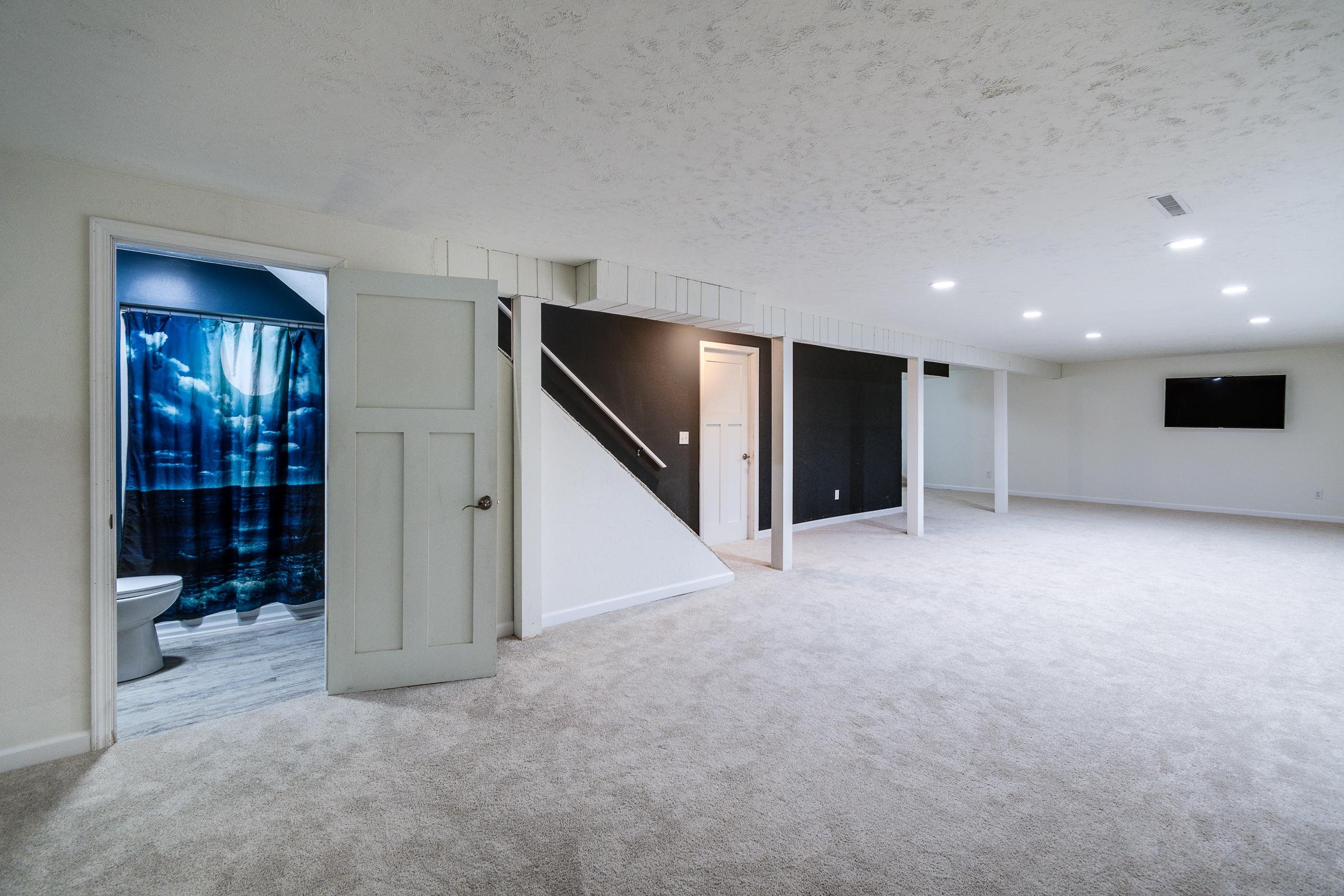 Carpeted basement rec room with support posts, stairs, a wall-mounted TV, and an adjoining bathroom.