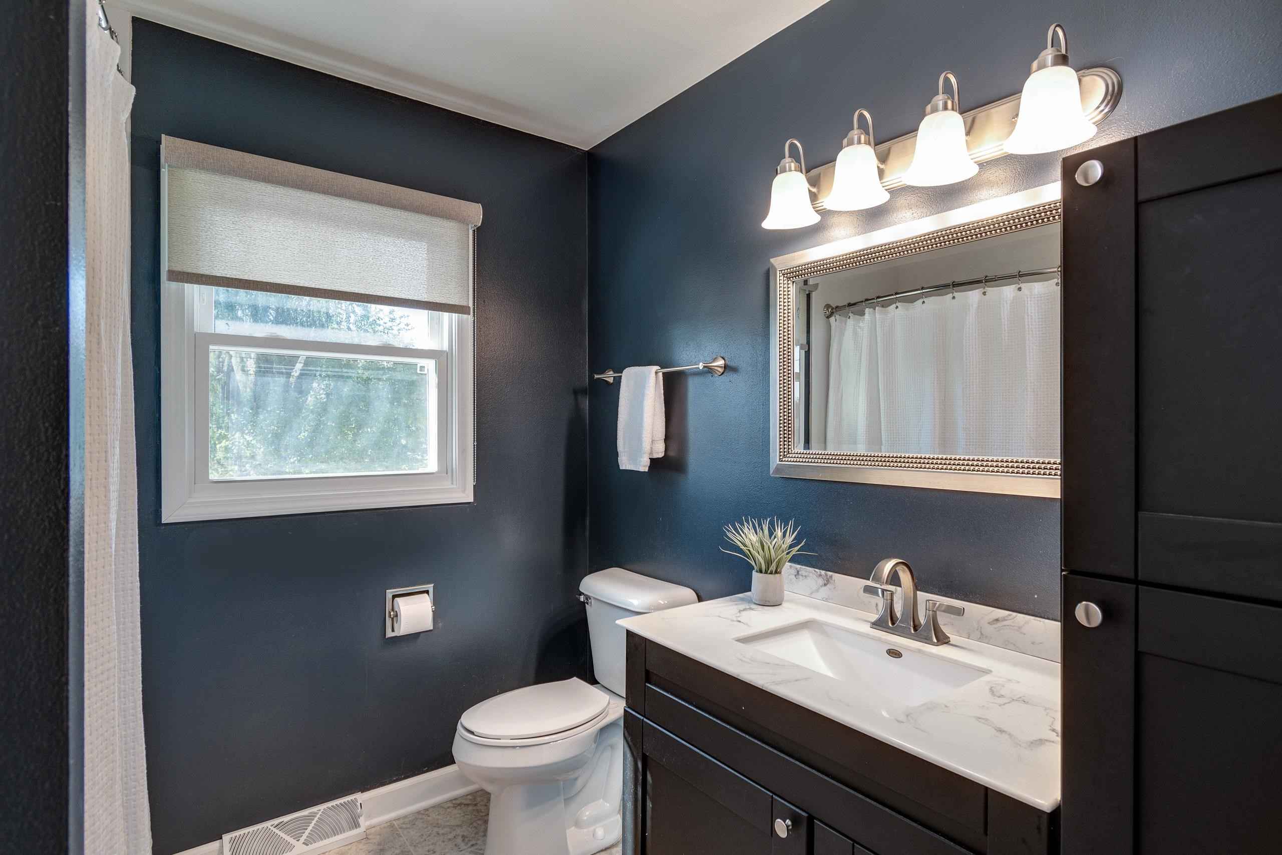 Bathroom with navy walls, a marble-look vanity, framed mirror, and window with roller shade.