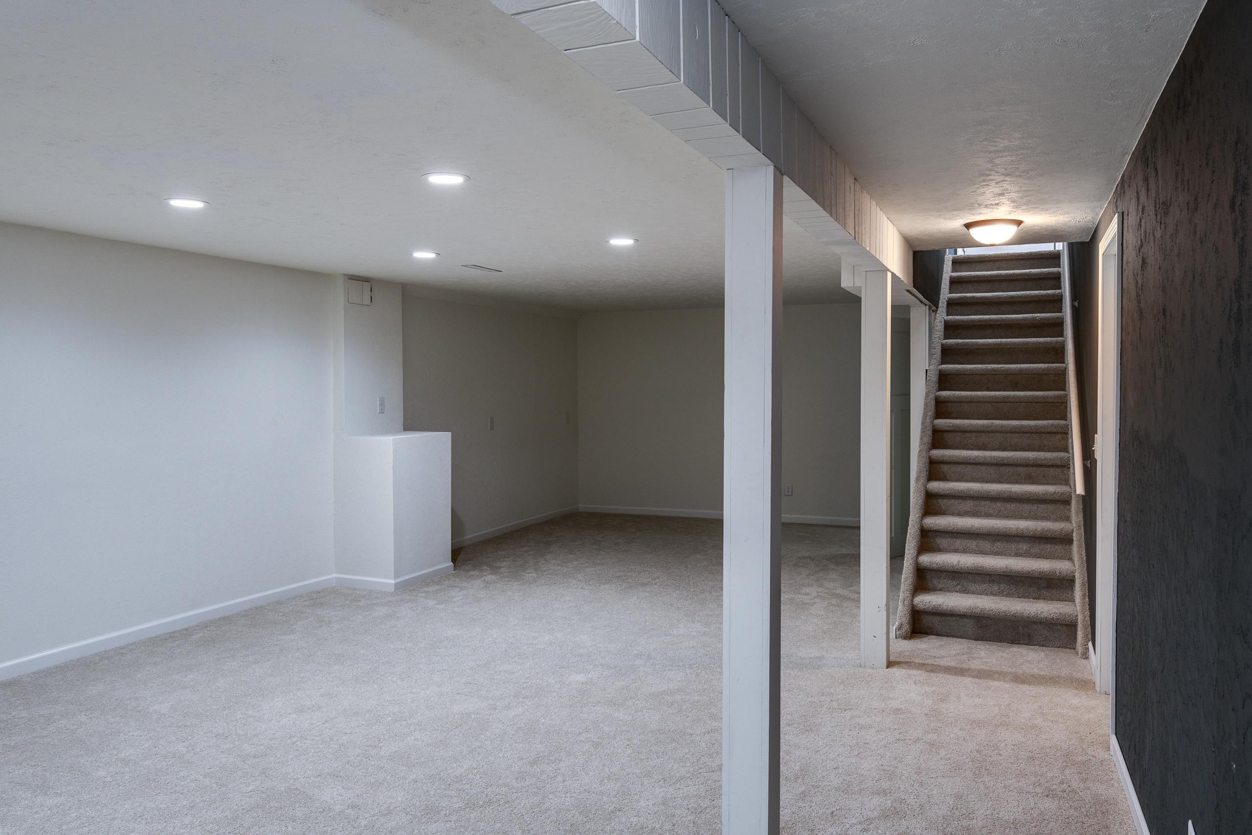 Empty carpeted basement with support columns and a staircase beside a dark accent wall.