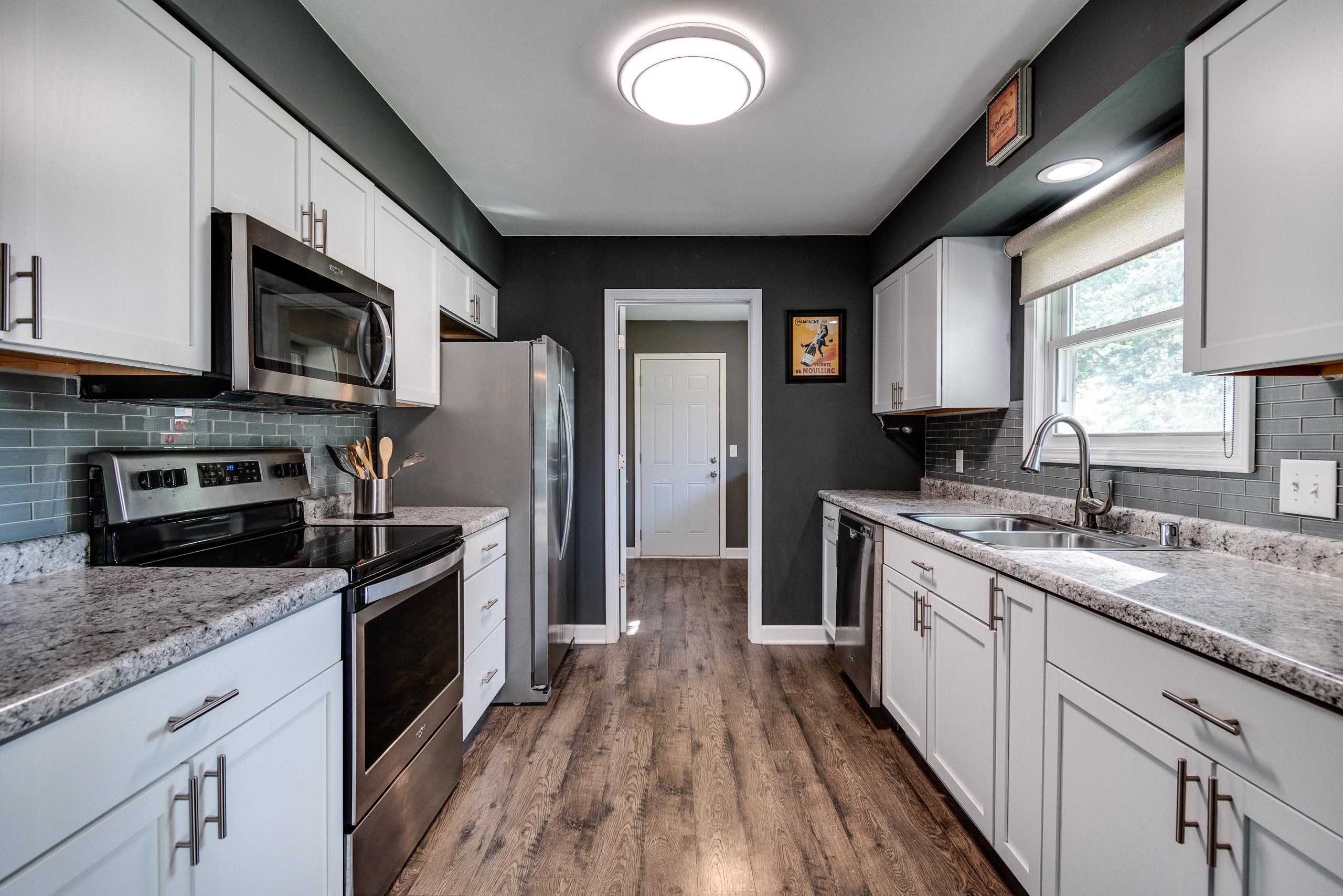 Galley kitchen with white cabinets, granite counters, stainless appliances, and a sink beneath a window.