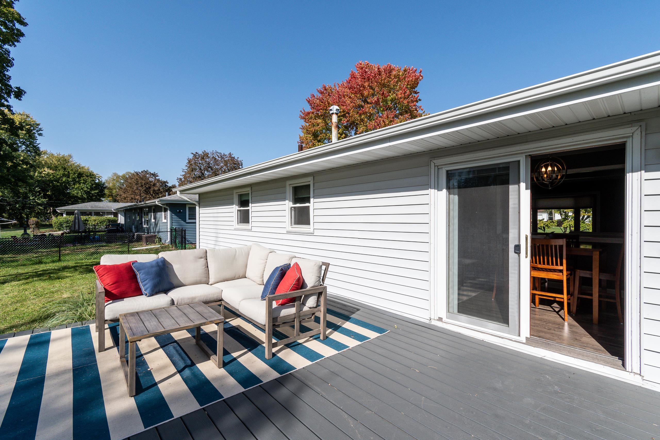 Gray-sided home with a furnished deck, striped rug, and open sliding door to the dining area.
