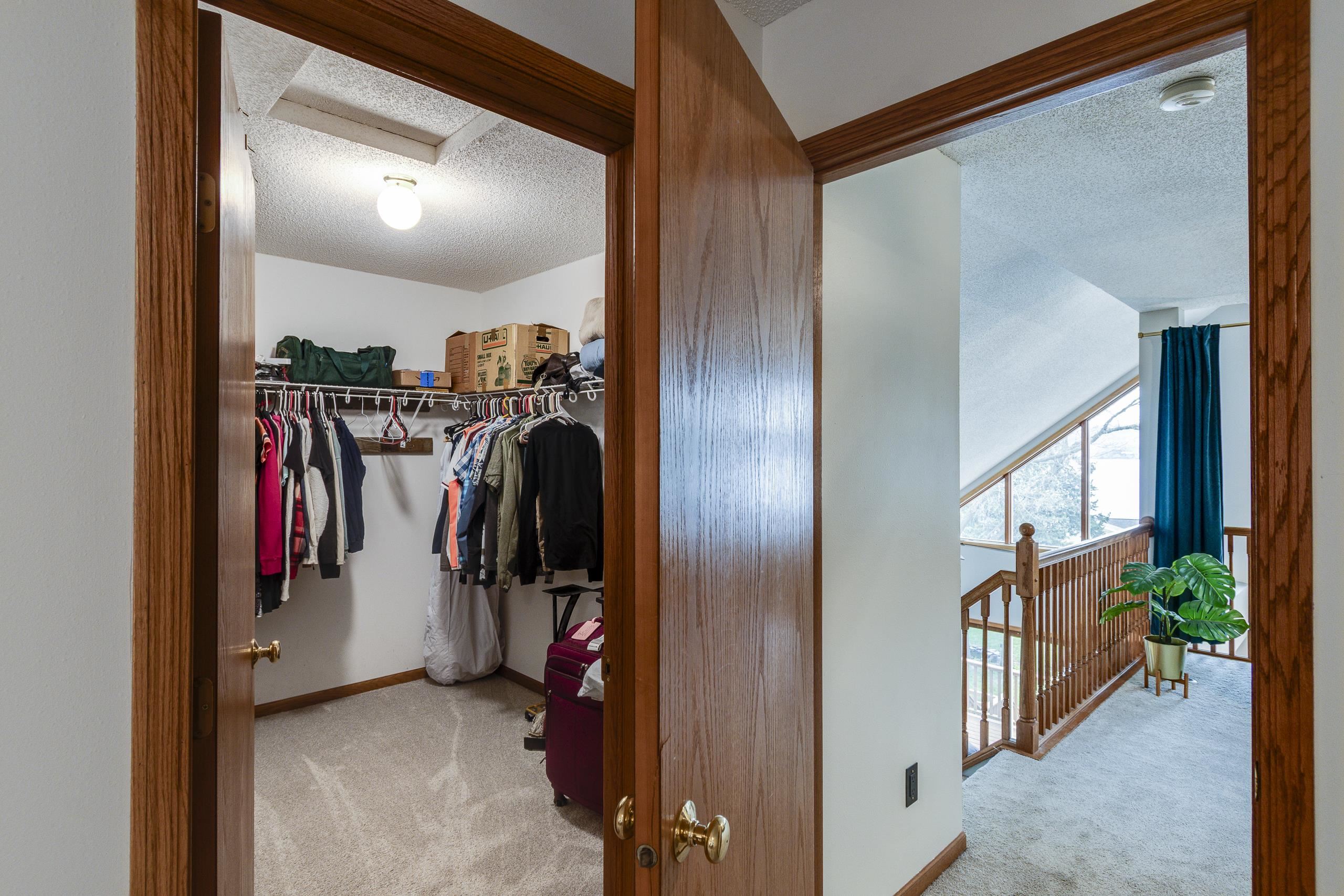 Walk-in closet beside an upstairs landing with a wooden railing, sloped window, and potted plant.