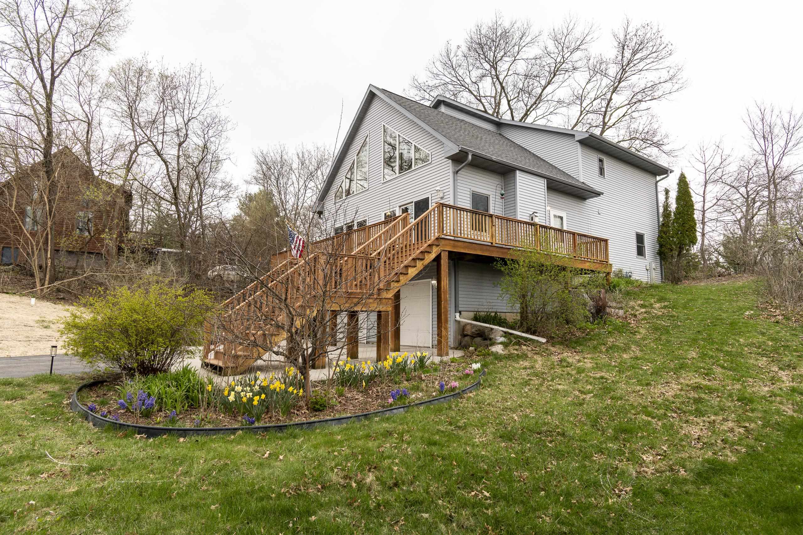 Gray-sided house with a large raised wooden deck, exterior stairs, and daffodils in the garden.