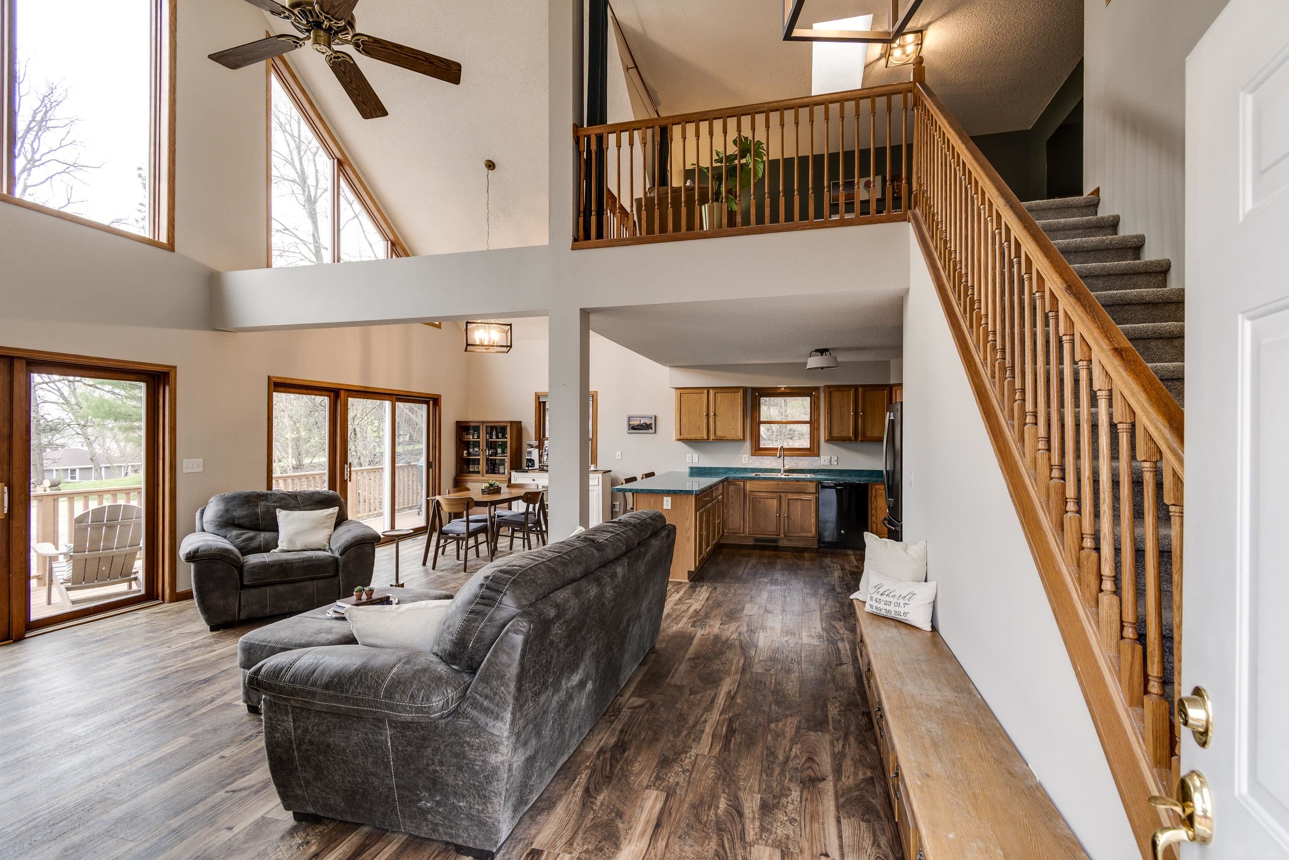 Open-plan living room with vaulted ceiling, gray sofas, tall windows, and a wooden staircase.