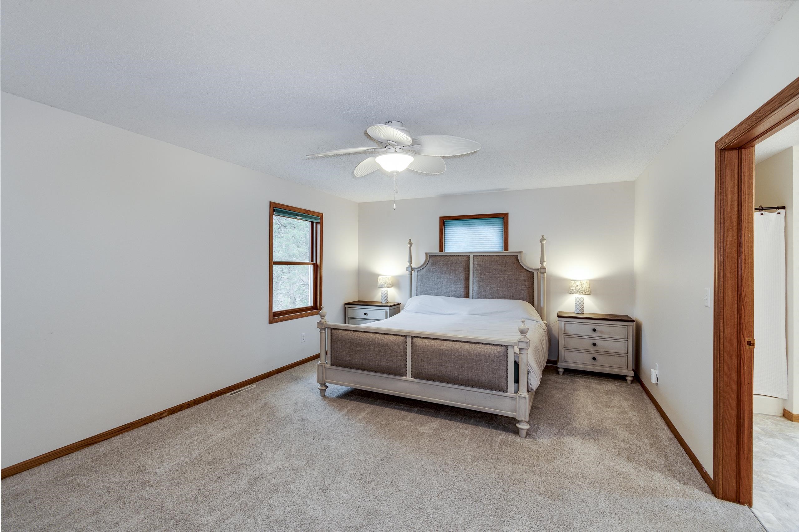 Furnished bedroom with a large bed, carpeted floor, wood-trimmed windows, and ceiling fan.