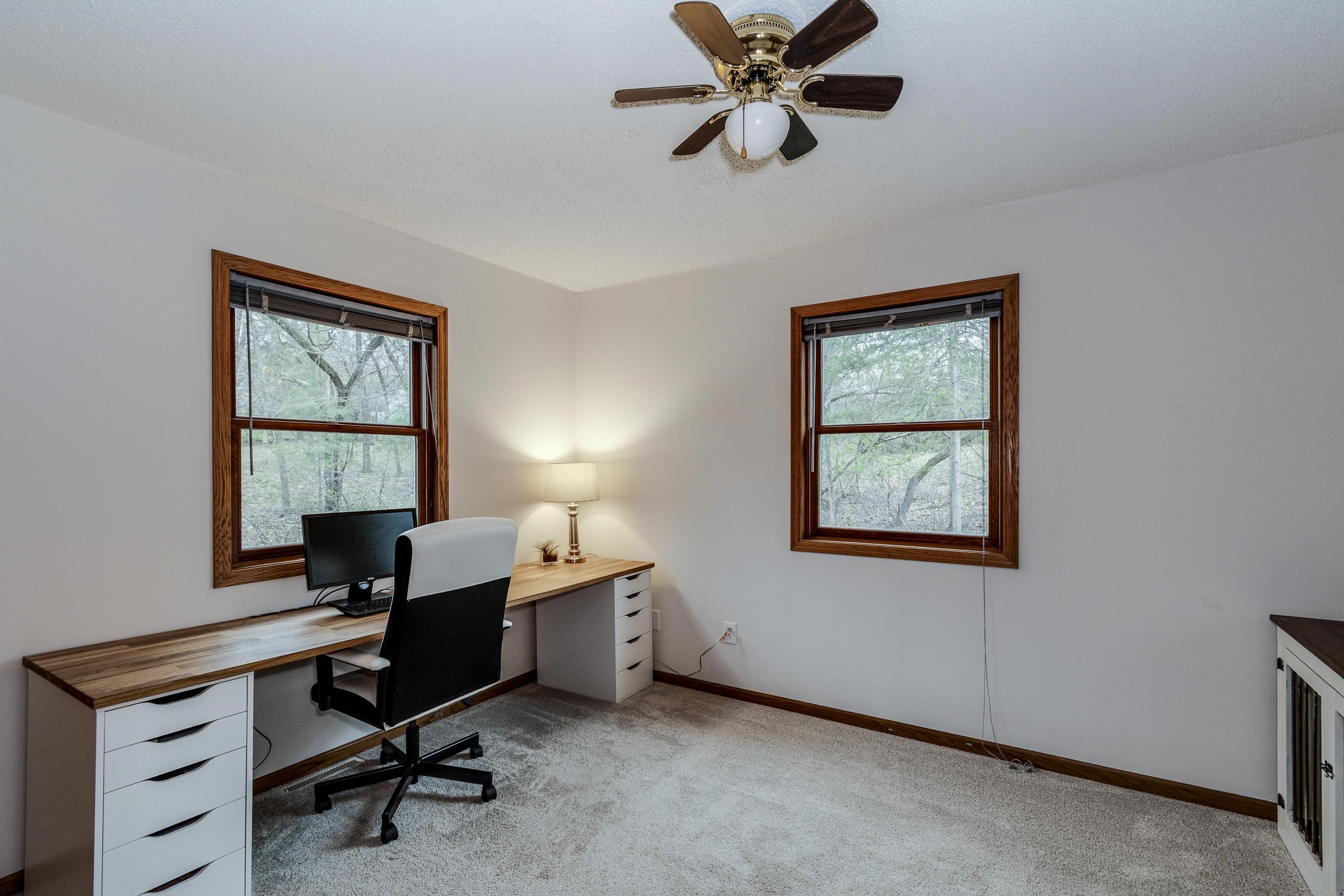 Home office with a long desk, computer, and two windows overlooking trees.