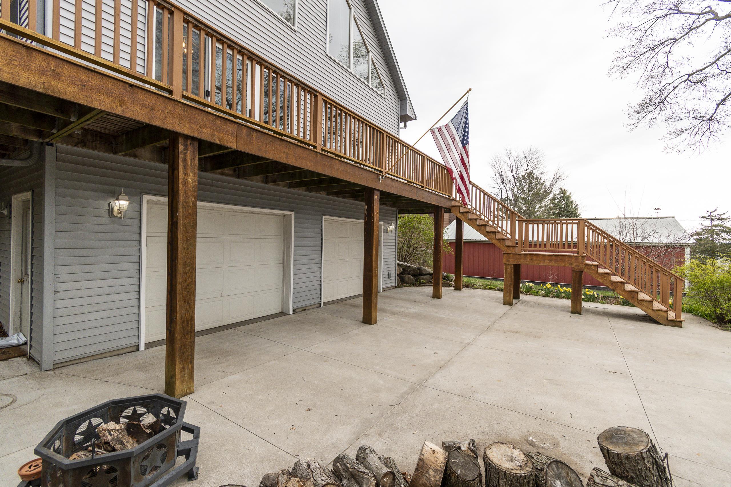 Home with an elevated wooden deck, long staircase, two garage doors, and a concrete patio with a fire pit.