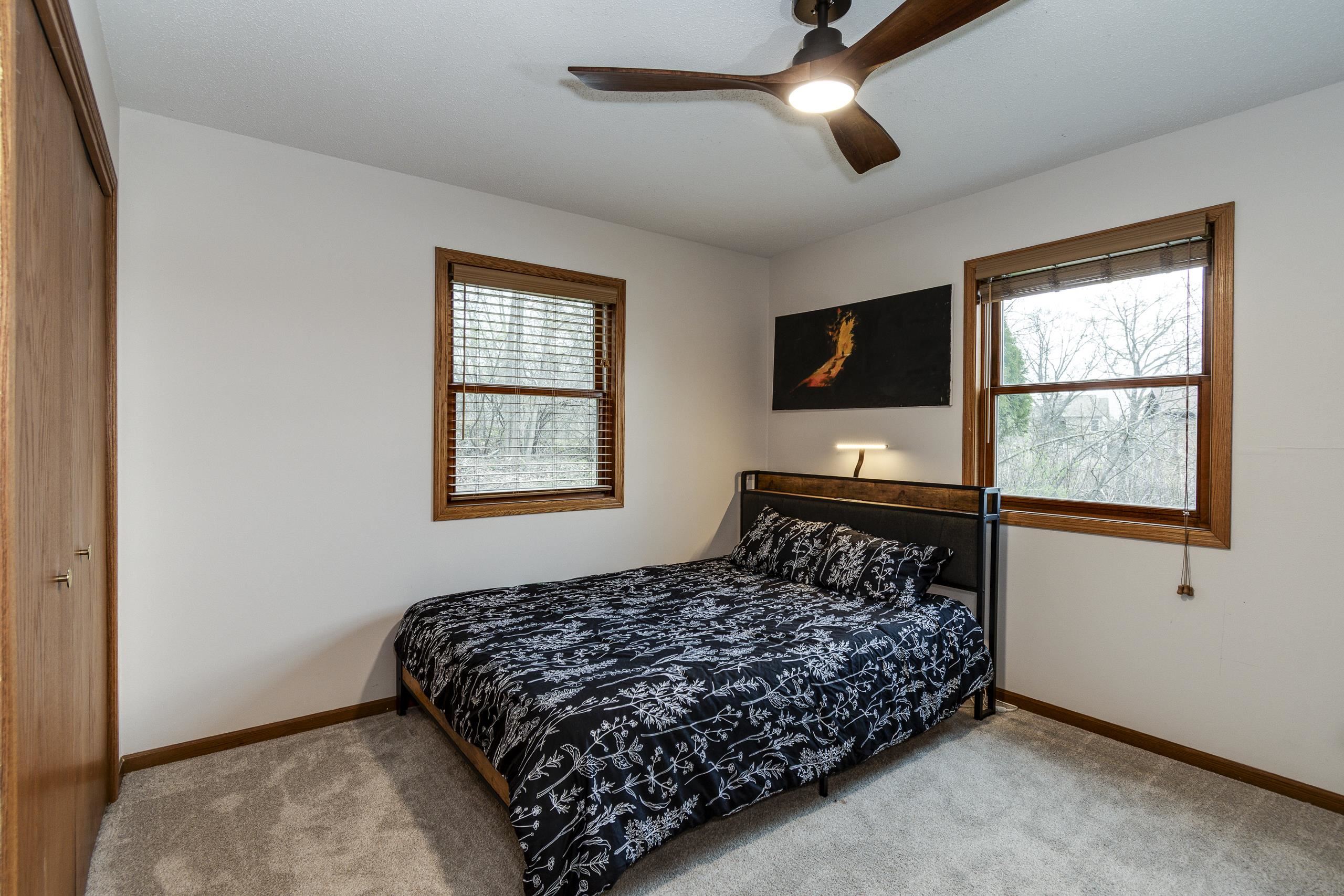 Bedroom with black floral bedding, wood-trimmed windows, beige carpet, and a ceiling fan.