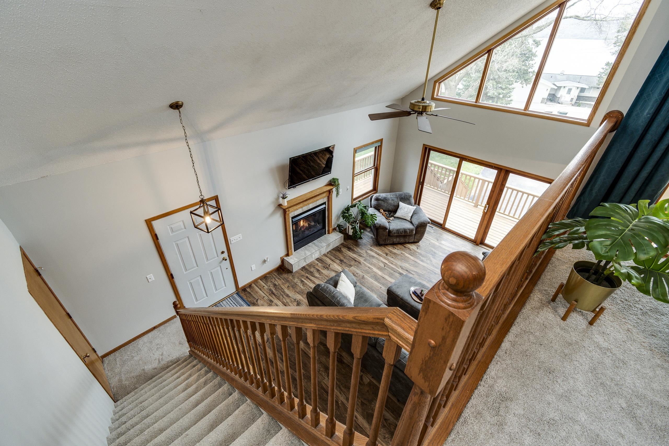 Open living room with vaulted ceiling, wood floors, fireplace, and large windows overlooking a deck.