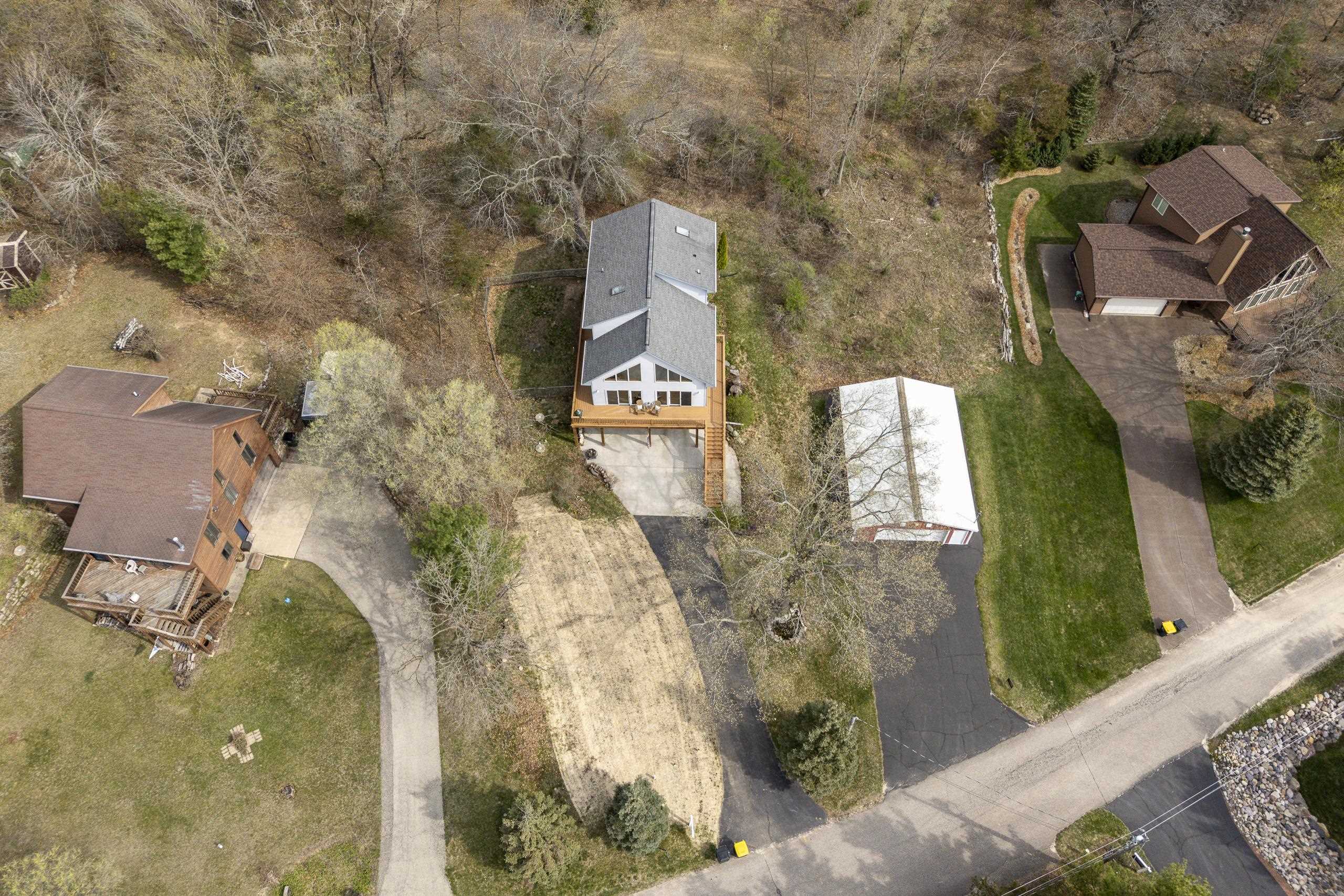 Aerial view of a home with a gray roof, deck, driveway, and wooded surroundings.