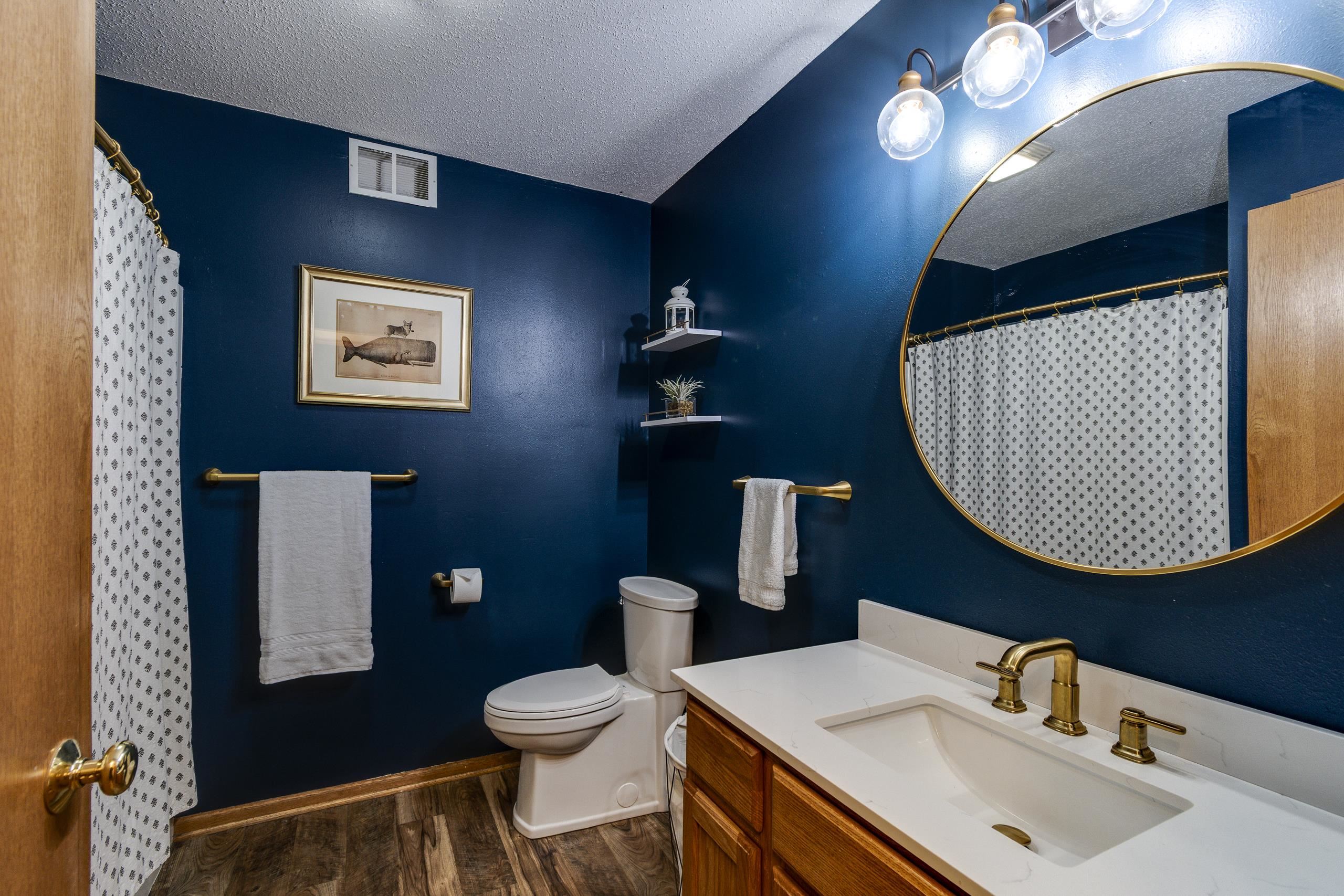 Bathroom with navy walls, a wood vanity, gold fixtures, and a large round mirror.