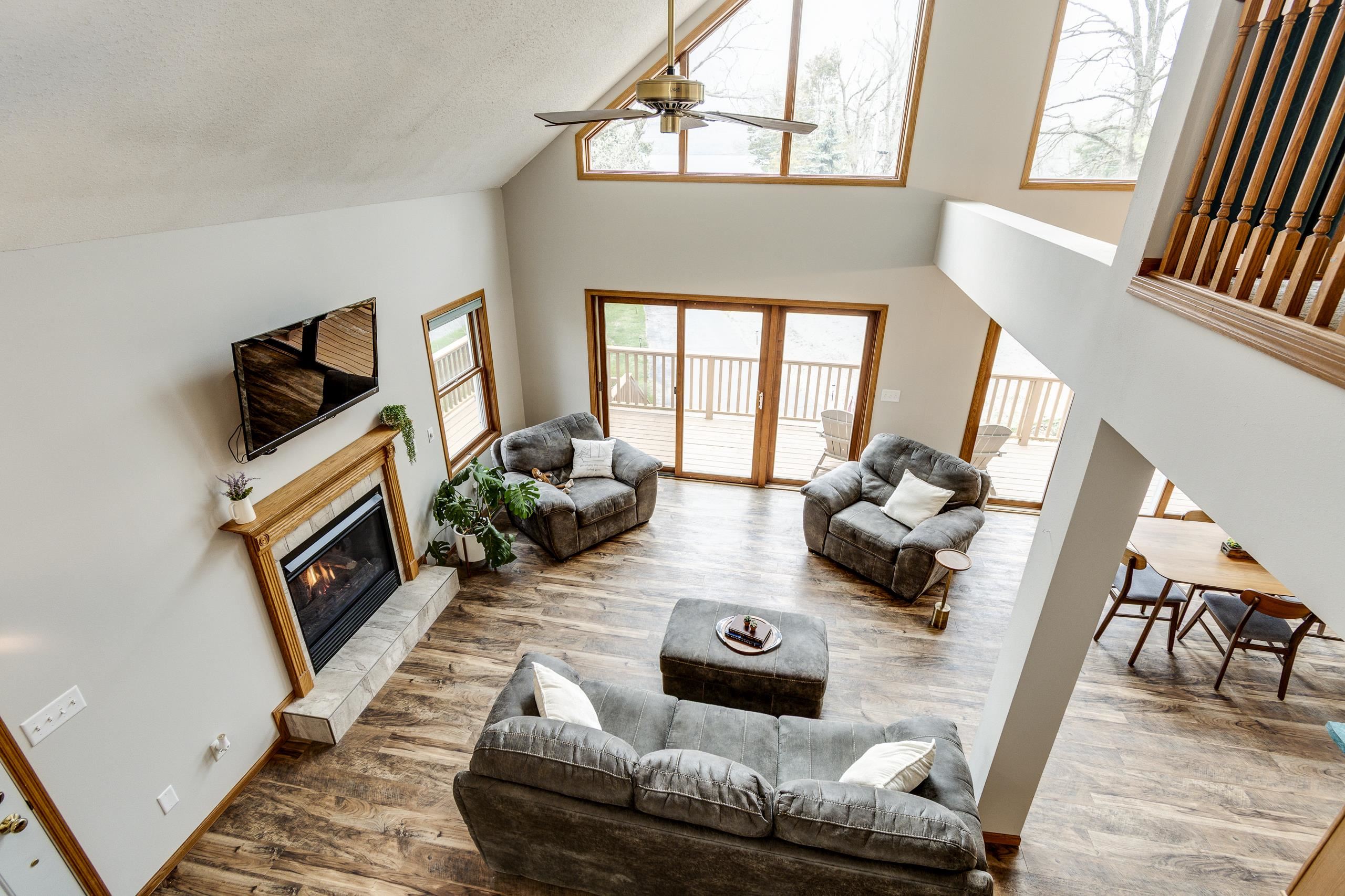 Vaulted living room with a fireplace, gray seating, and large windows opening onto a deck.