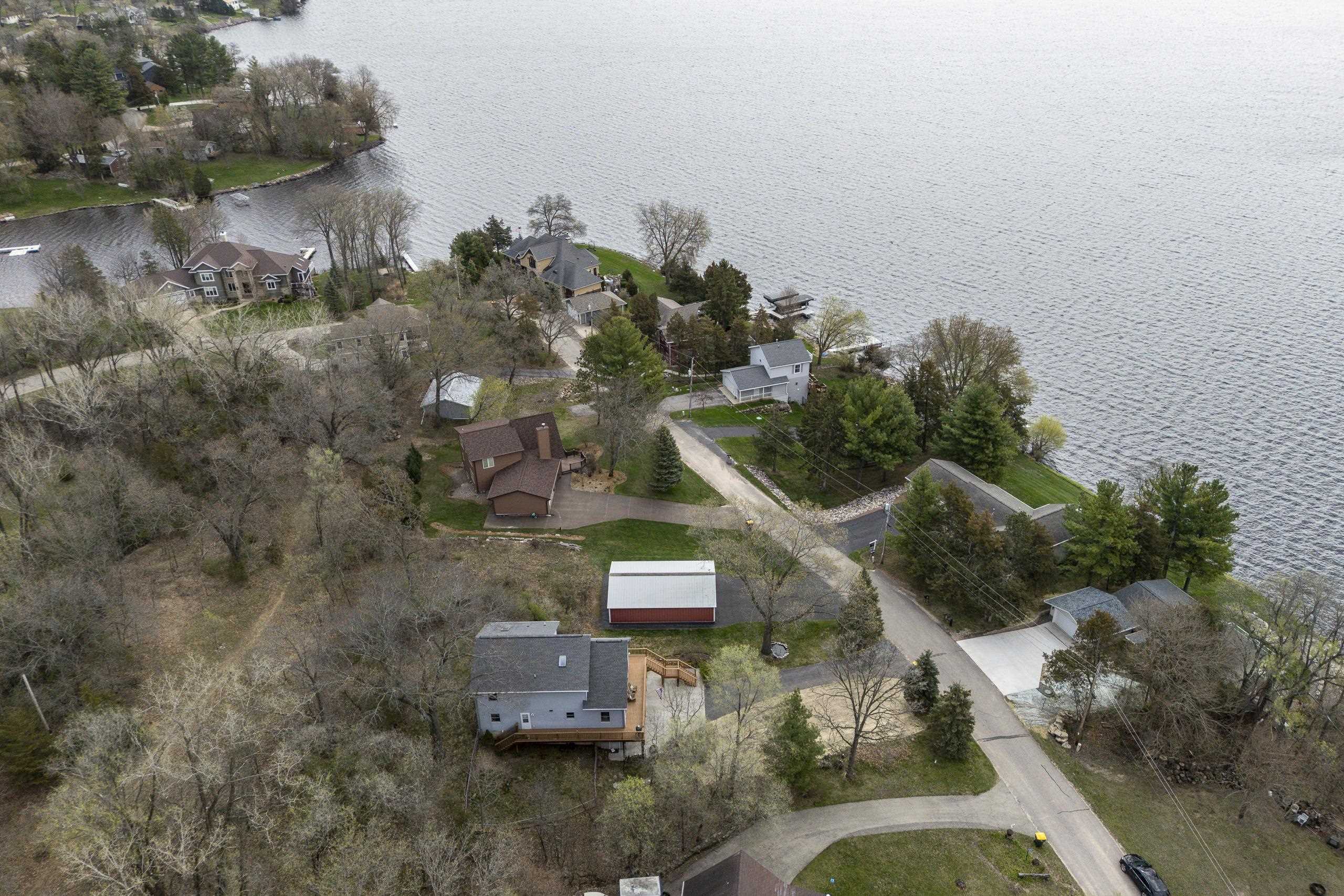 Aerial view of a gray lakefront home with a deck, wooded lot, and red outbuilding.