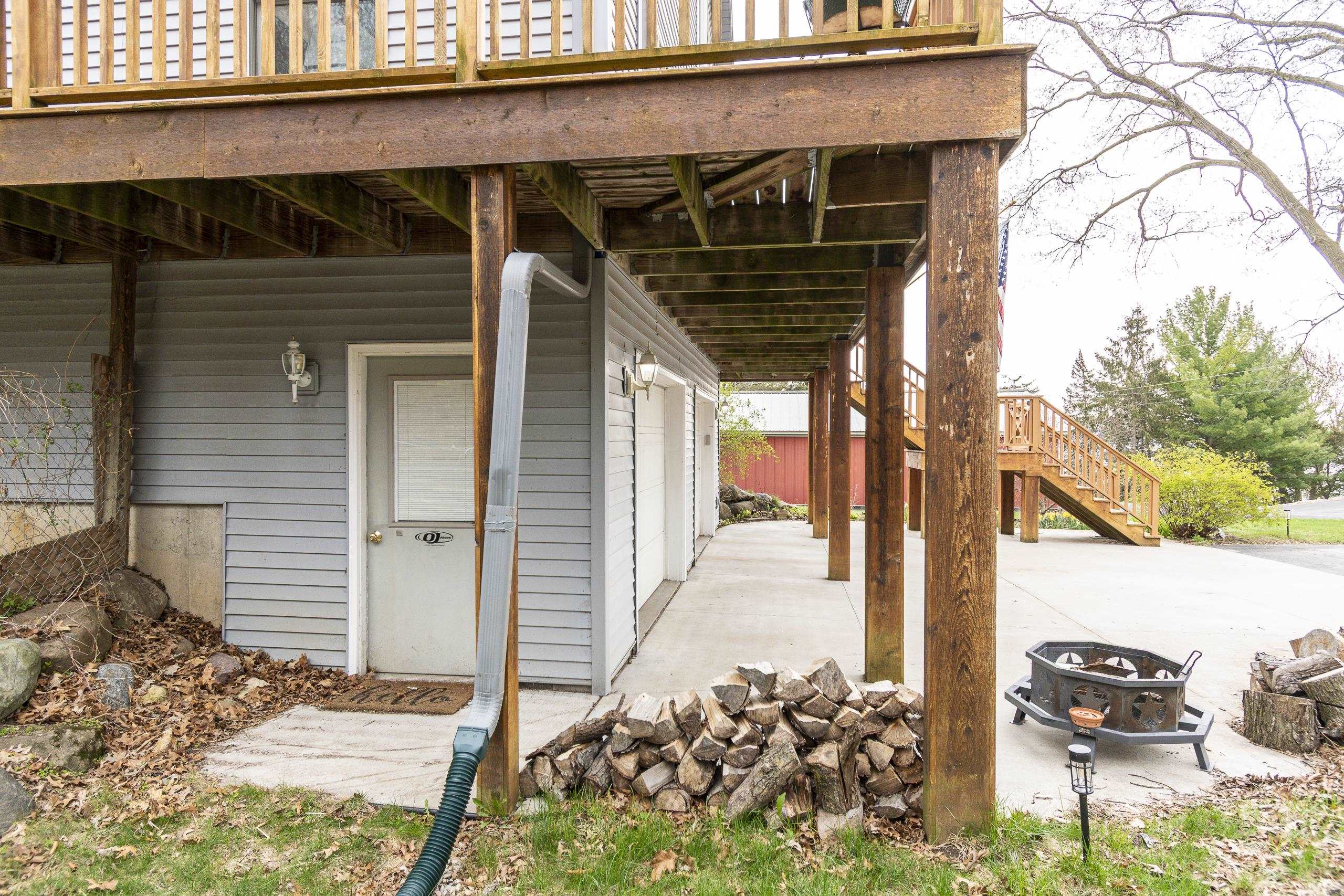 Covered concrete patio beneath a raised wooden deck, with timber posts, stairs, a fire pit, and stacked firewood.