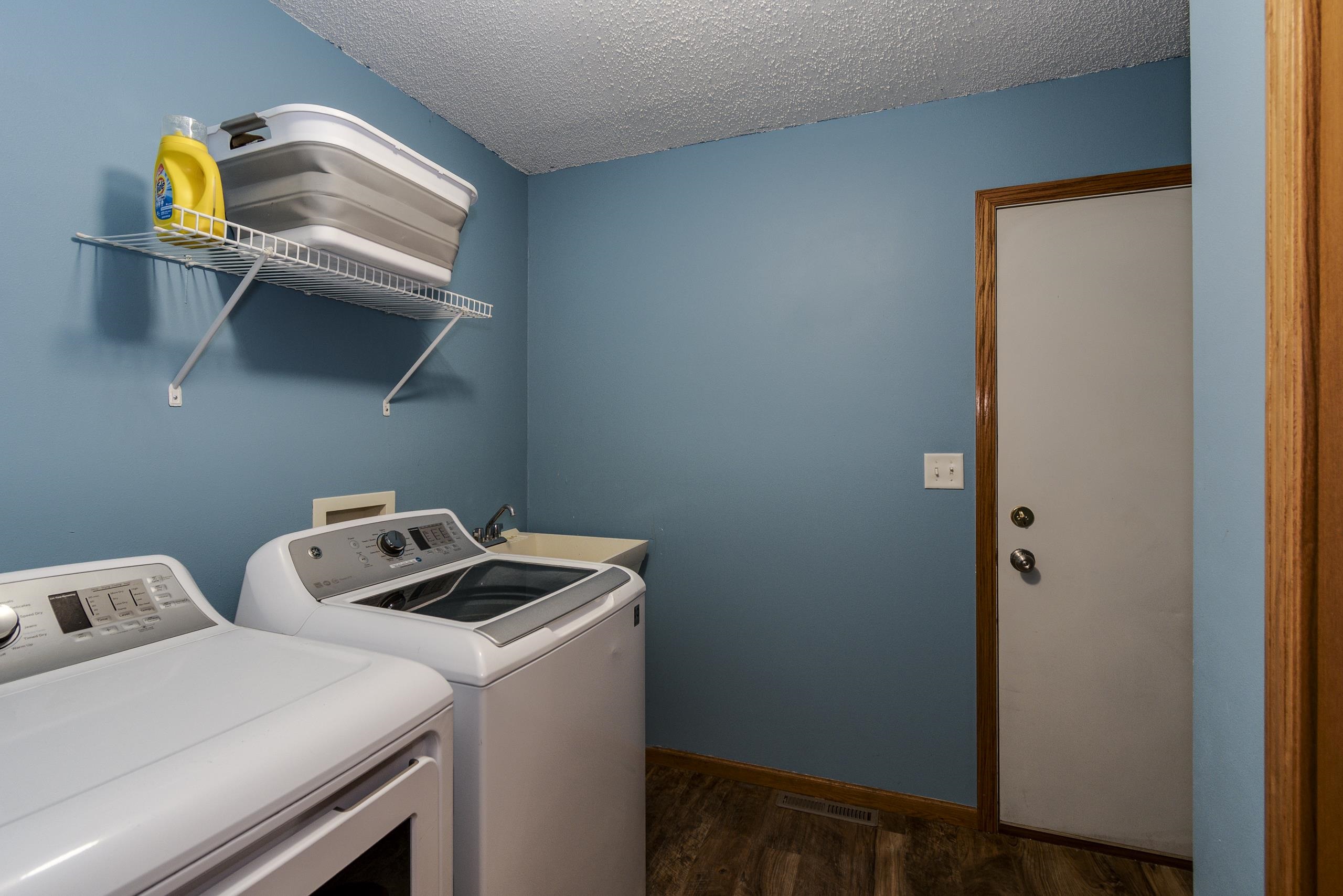 Laundry room with white washer and dryer, utility sink, and wire shelf against blue walls.