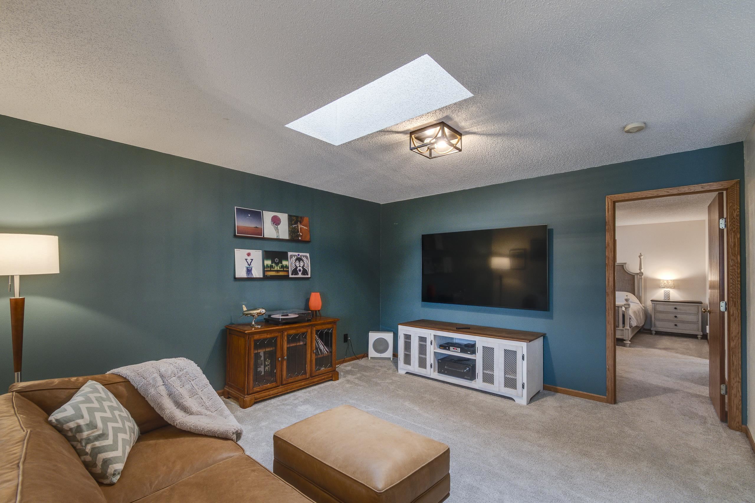 Living room with teal walls, leather sectional, wall-mounted TV, and skylight; bedroom visible through the doorway.