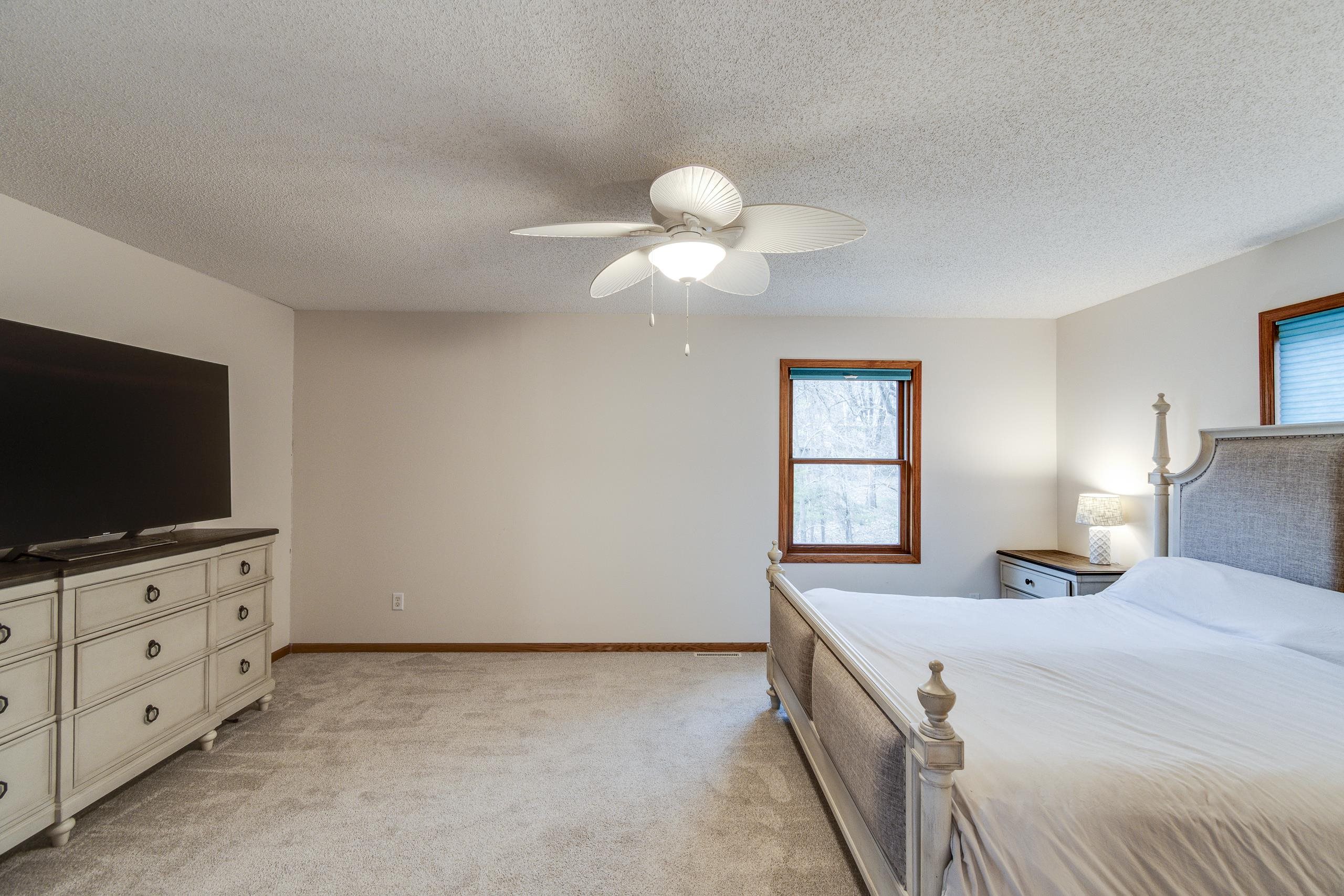 Furnished bedroom with a large bed, beige carpet, ceiling fan, and wood-trimmed window overlooking trees.