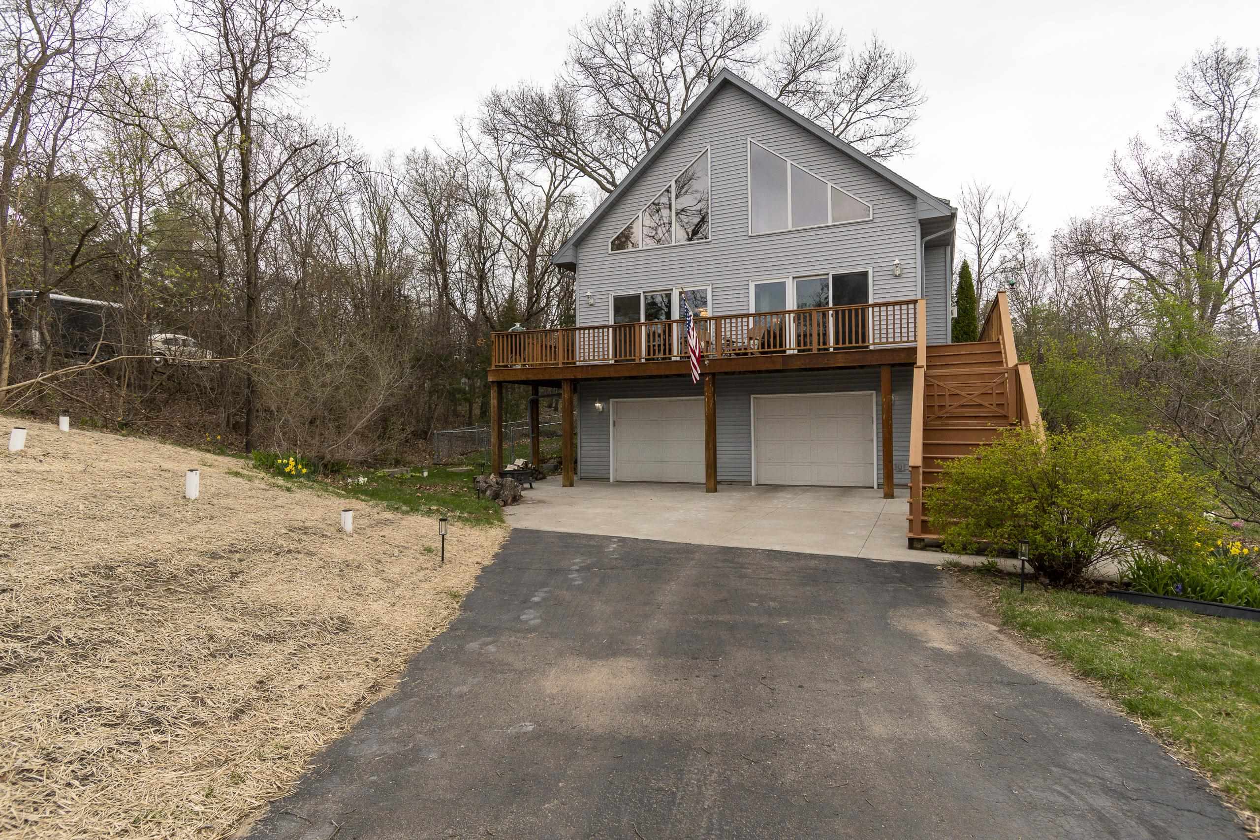 Gray gabled home with a raised wooden deck, exterior stairs, and two-car garage beside a wooded driveway.