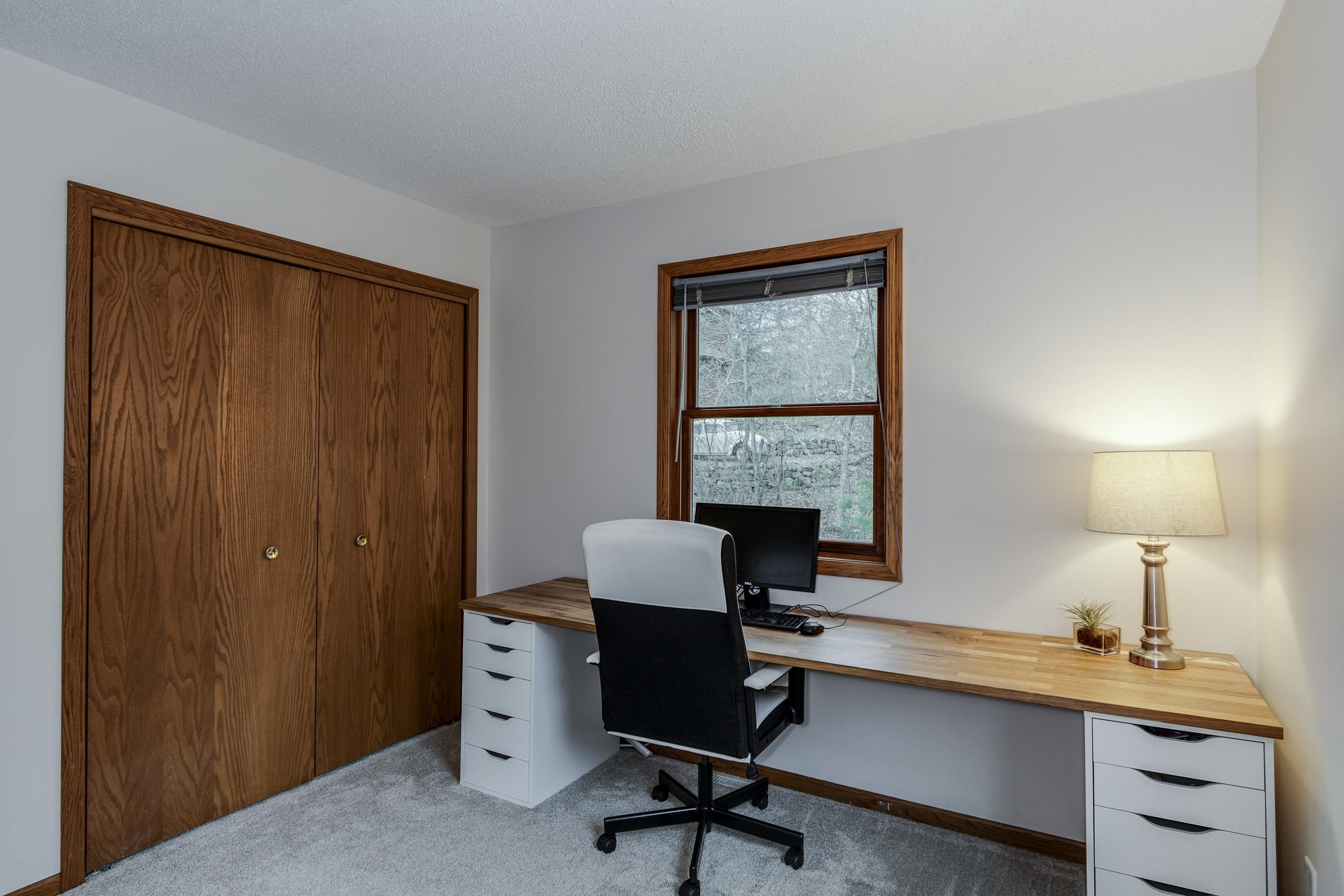 Home office with a long wood desk, black chair, wood-trimmed window, and large wood closet.