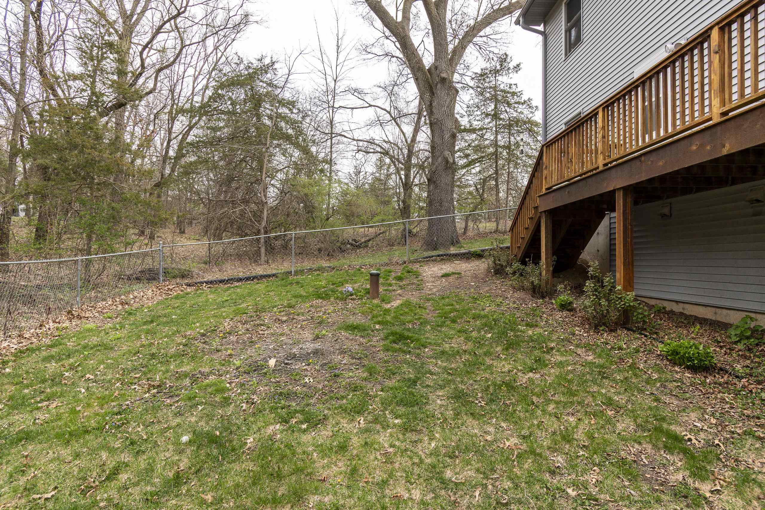 Grassy backyard beside a gray home, with a raised wooden deck, chain-link fence, and mature trees.