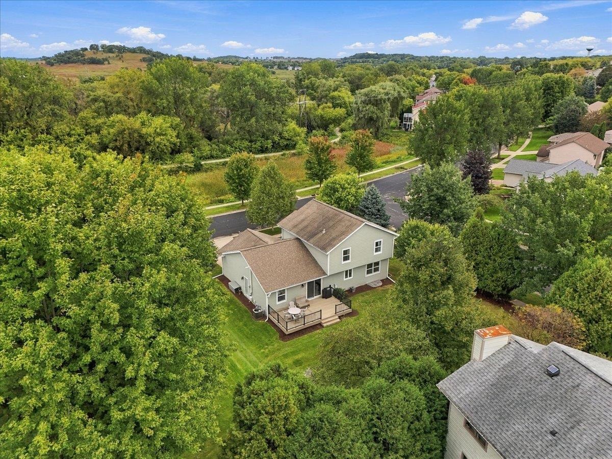 Aerial view of a two-story home with a deck, broad green yard, and wooded hills beyond.